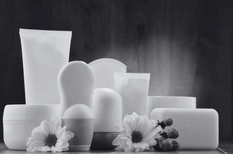 A collection of white cosmetic containers, including tubes, jars, and bottles, are arranged with white flowers and berries against a dark, textured background.