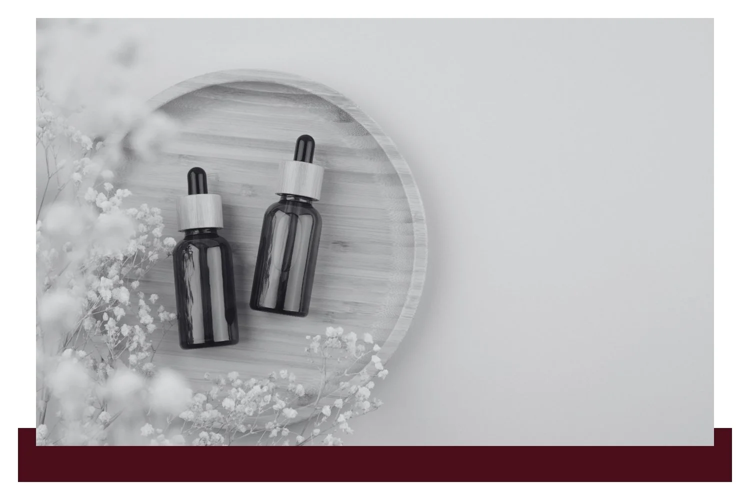 Two dark glass essential-oil bottles with dropper tops rest on a round wooden tray, surrounded by soft white mist and small white flowers against a light background.