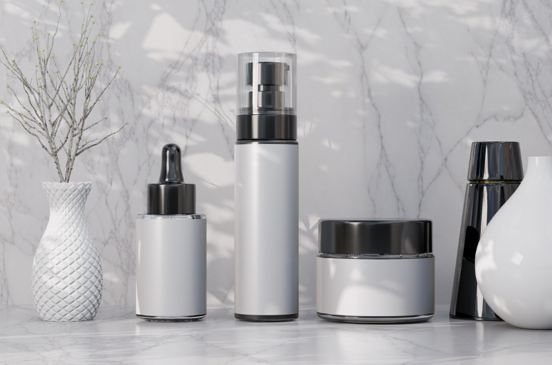 A collection of white and black cosmetic bottles and jars arranged on a marble surface with a decorative white vase and dried branches to the left.