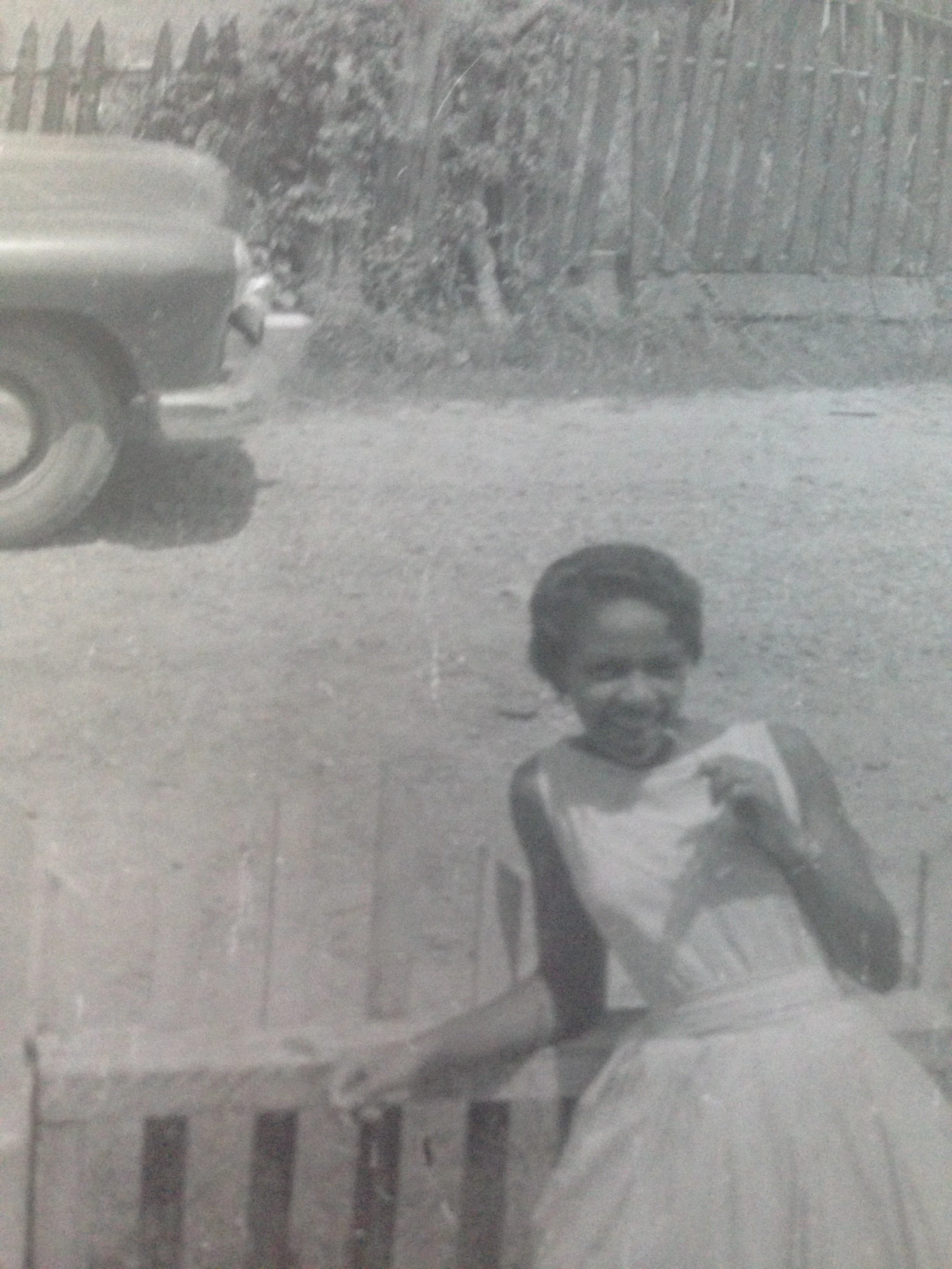 Photo of author's mom. Black woman wearing a white sleeveless dress with a pair of glasses, standing against a wooden fence.