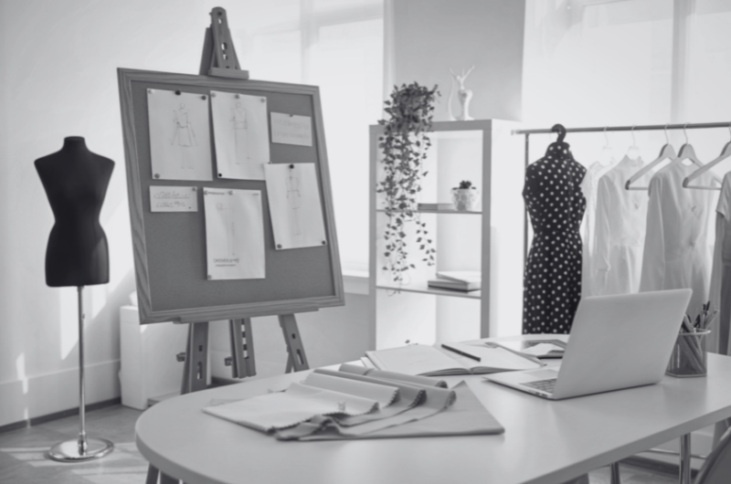A sewing studio with a dress form, sketches on a corkboard, fabric on a table, and clothes on a rack.