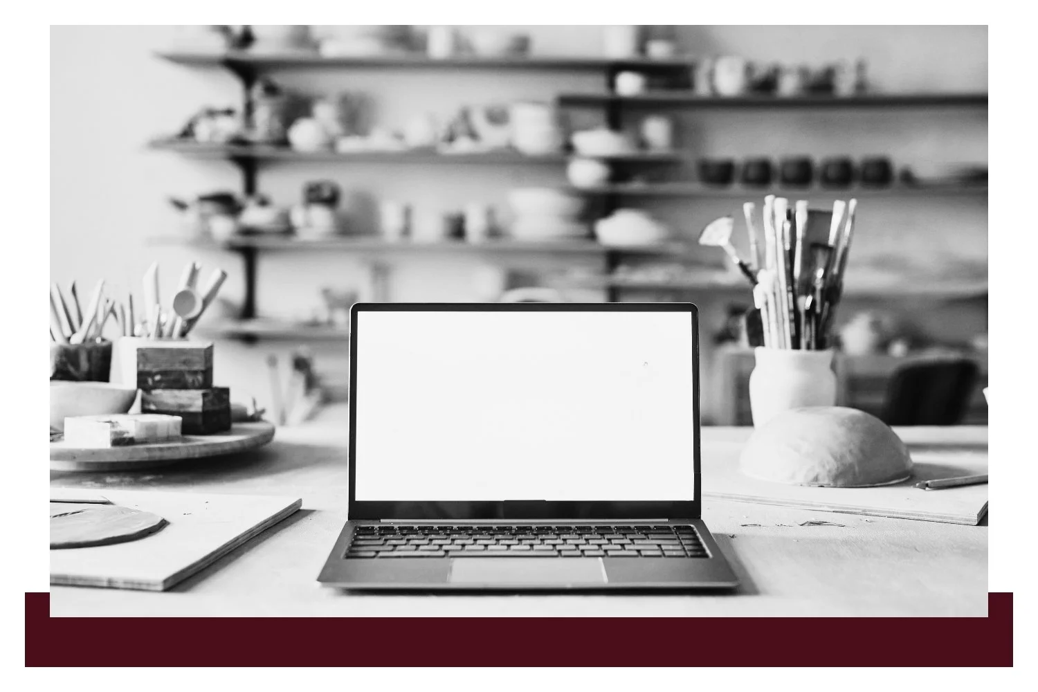 An open laptop with a blank white screen sits on a desk with art supplies nearby, including paintbrushes in a cup and stationery, while shelves of tools blur in the background.