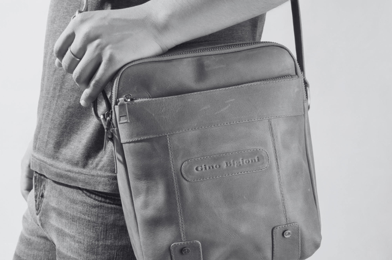 A person wearing a grey t-shirt and jeans, carrying a grey crossbody bag with the "Gino Biglioni" logo embossed on the front.
