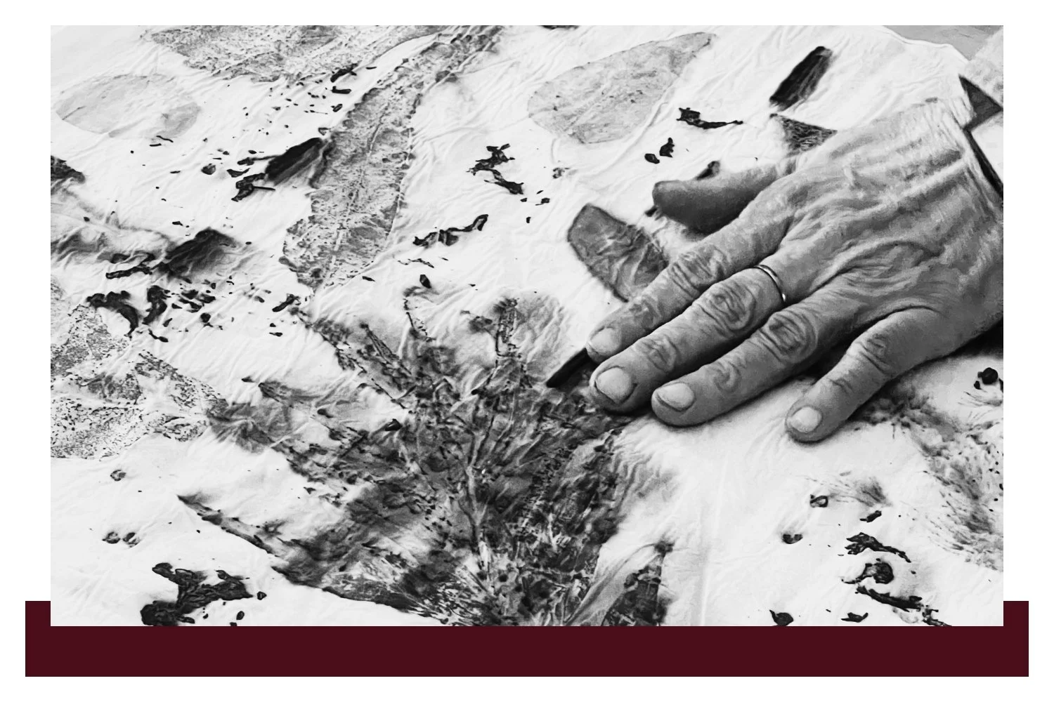 Gloved hands in the right foreground hold a pencil over a snowy, war-damaged landscape, while scattered debris and dark smudges spread across the ground in the background.