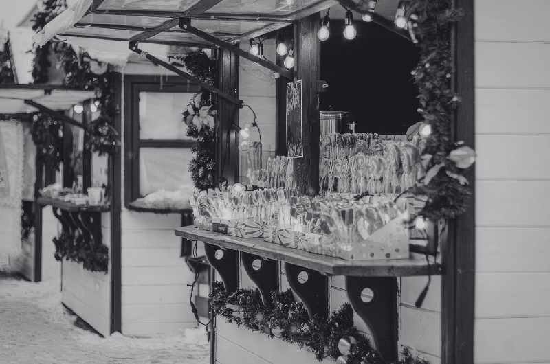 A black and white image of a festive outdoor market stall decorated with garlands and lights, displaying numerous items on shelves.
