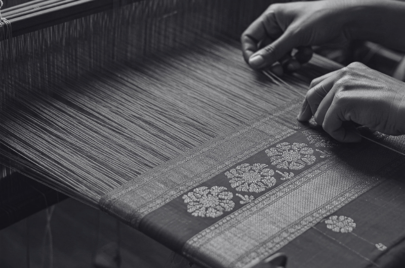 A close-up, black and white shot of hands working on a loom, weaving a patterned fabric.