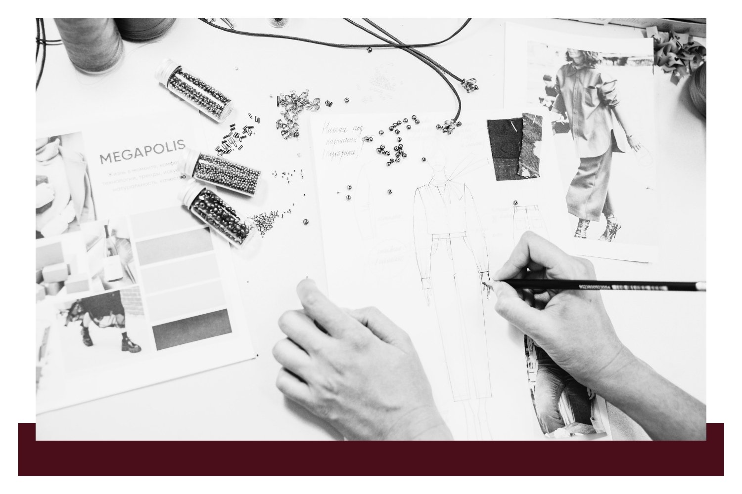 Hands drawing on a white desk covered with architectural sketches, diagrams, and printed photos; a person holds a pencil over the plan while other documents and wires lie nearby.