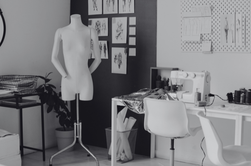 A white mannequin stands in a room with a sewing machine, spools of thread, and fashion sketches on the wall.