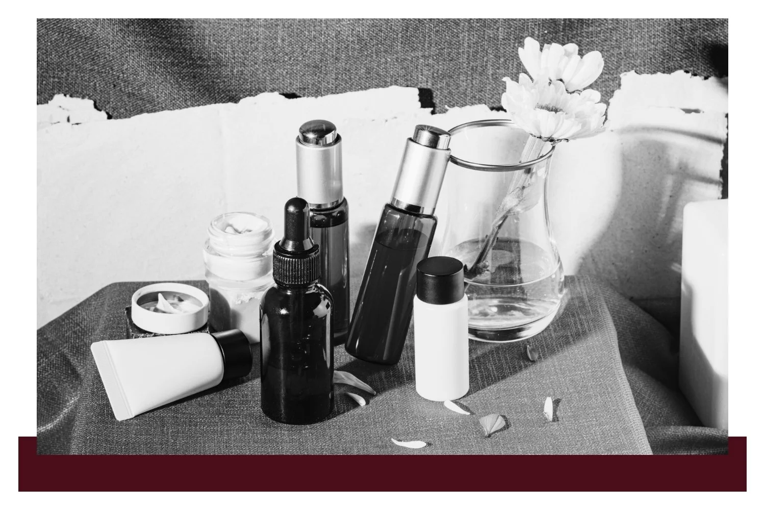 Black-and-white still life of skincare bottles and containers on a table, with a glass beaker holding water and a small white flower in the background.