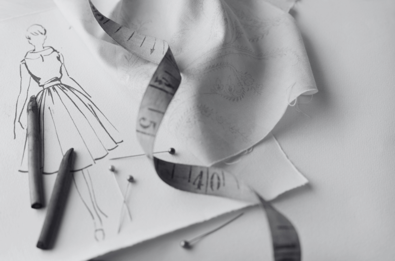 A fashion sketch of a dress with a measuring tape, pins, and pencils on a white surface.