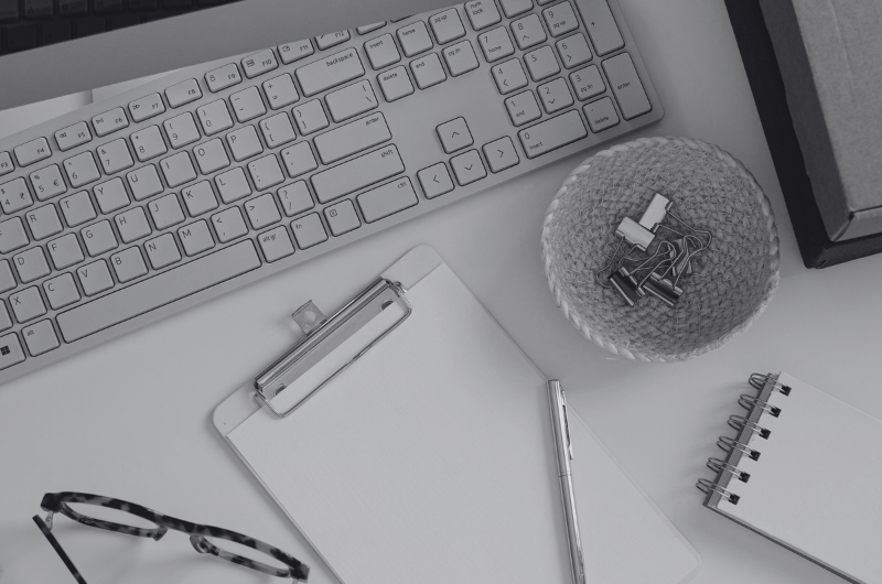 A flat lay of a desk with a keyboard, clipboard, pen, eyeglasses, a basket of paper clips, a notebook, and a box.