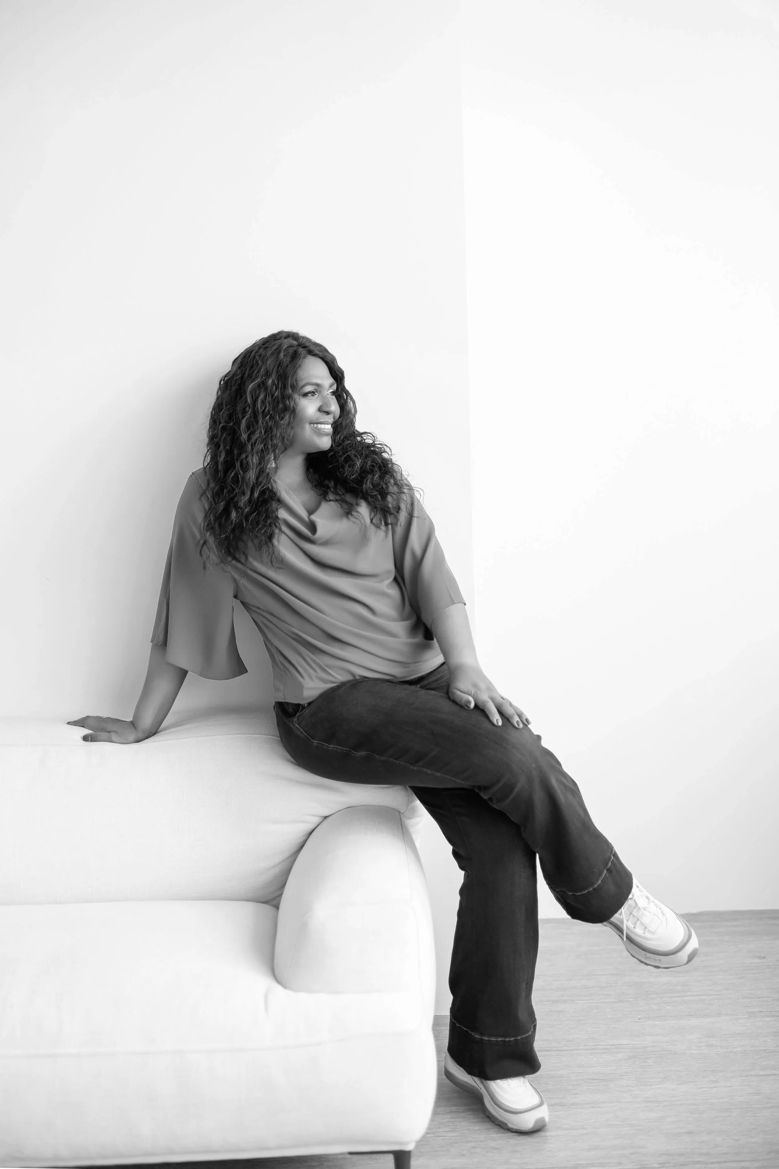 C. Nicole Gaither, founder of Cloth & Code Legal, seated on a white sofa against a white wall, looking off to the right