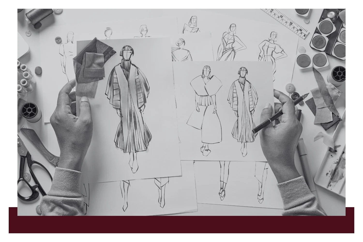 Hands pin and adjust a pattern paper showing several dress and garment sketches with labels, surrounded by sewing tools, scissors, fabric swatches, and rulers on a worktable.