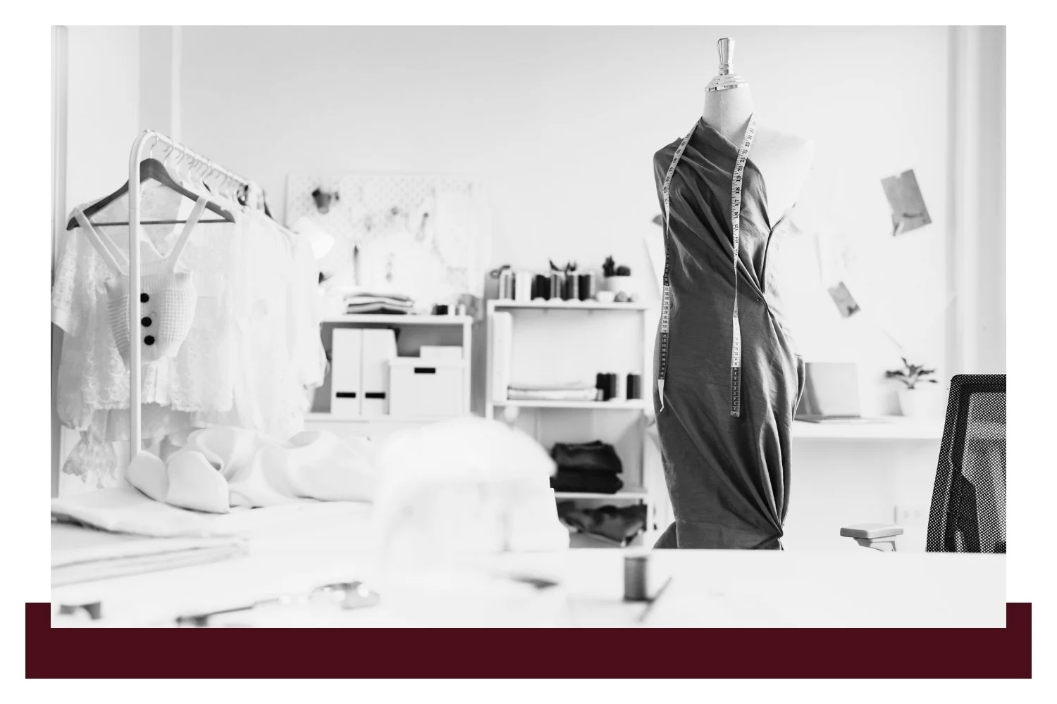 Black-and-white studio scene with a dress form wearing a dark garment and a sewing table with hanging fabrics, tools, and boxes in the background.