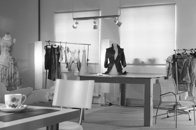 A studio space with clothing racks, mannequins displaying garments, and a table with chairs. A jacket is displayed on a mannequin on the table.
