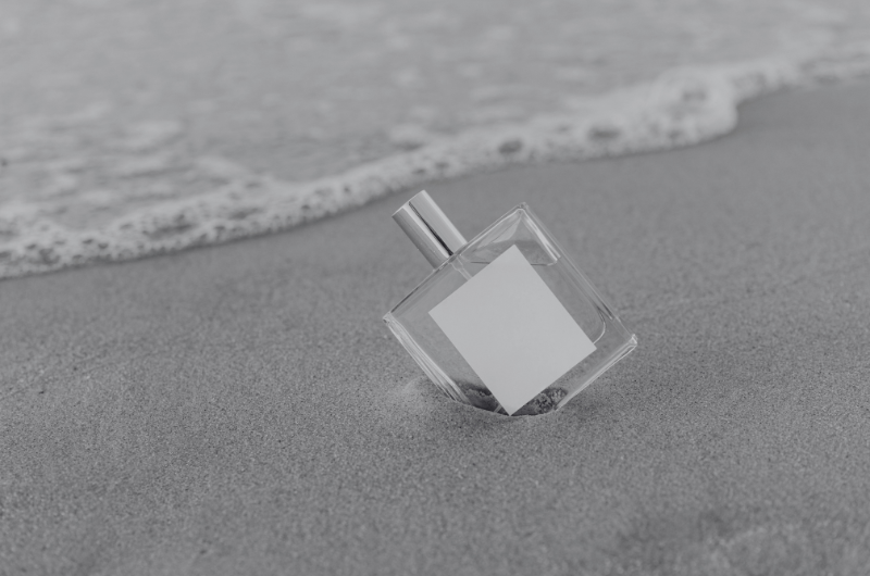 A clear glass perfume bottle with a silver cap rests on a sandy beach, with gentle waves lapping at the shore.