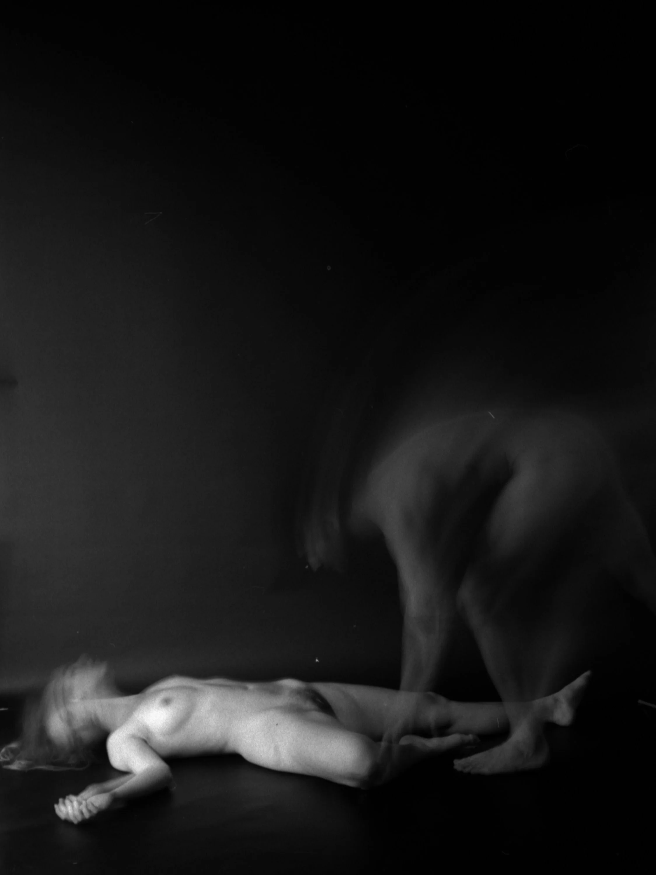 Black and white image of a naked woman lying on her side with her back facing upward against a dark background.