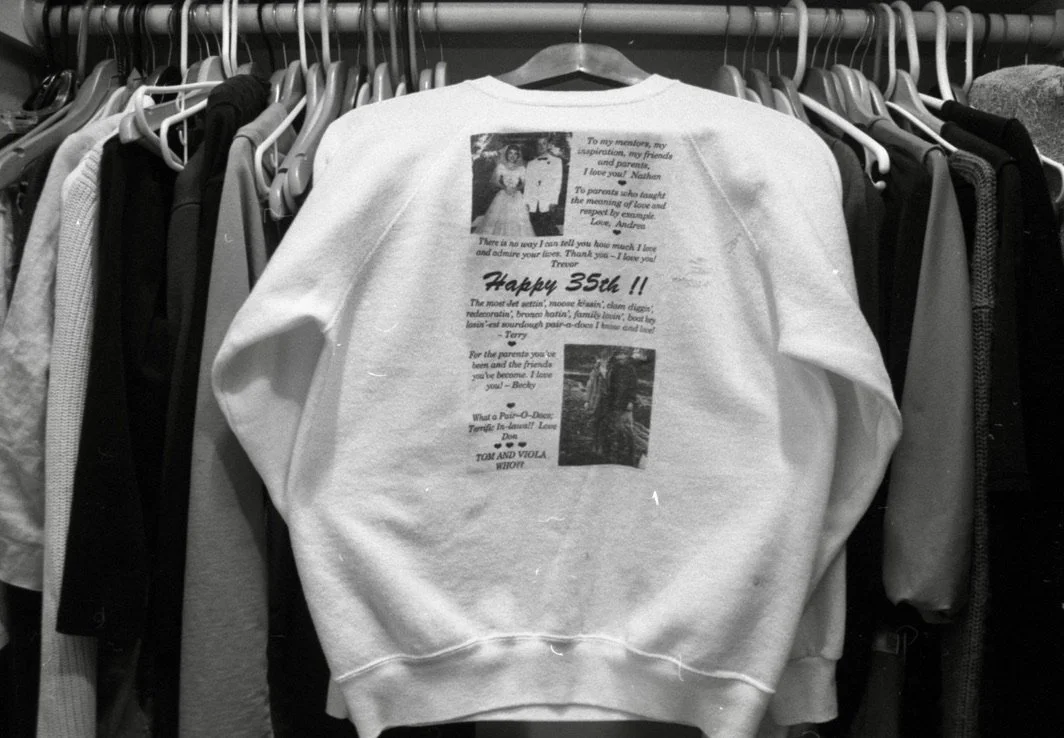 White sweatshirt hanging on a clothing rack with printed text and images on the back, surrounded by other garments.