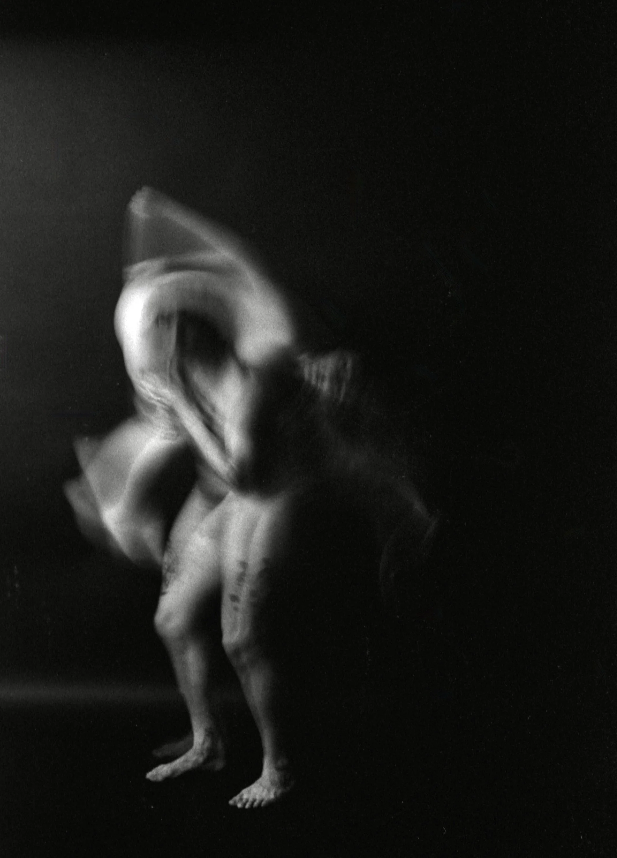 A black-and-white photograph of a humanoid figure with an elongated head, standing against a dark background. The figure appears to have a distorted or abstract form with exaggerated features.