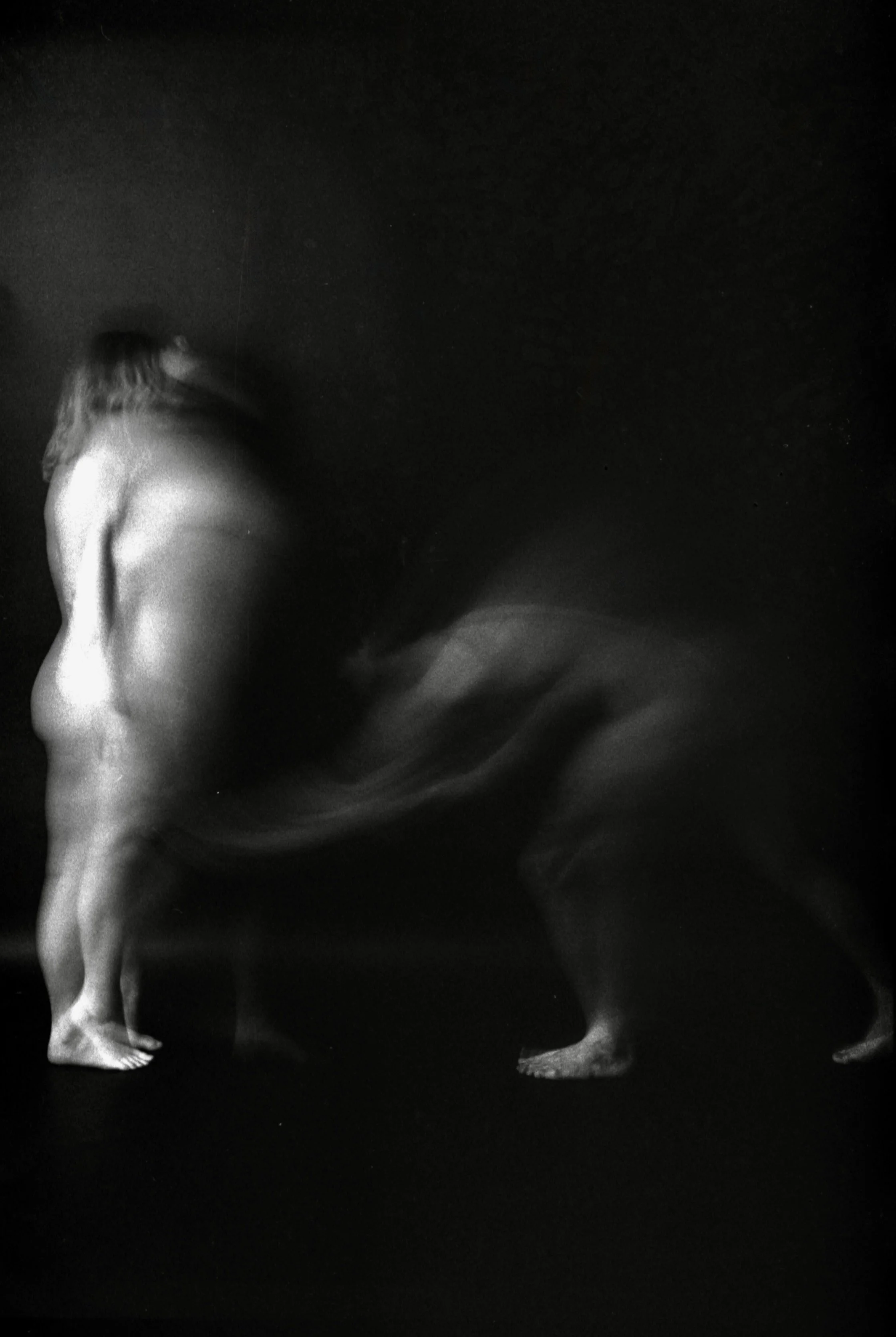 Black and white photograph of a naked woman with long hair standing on a dark background.