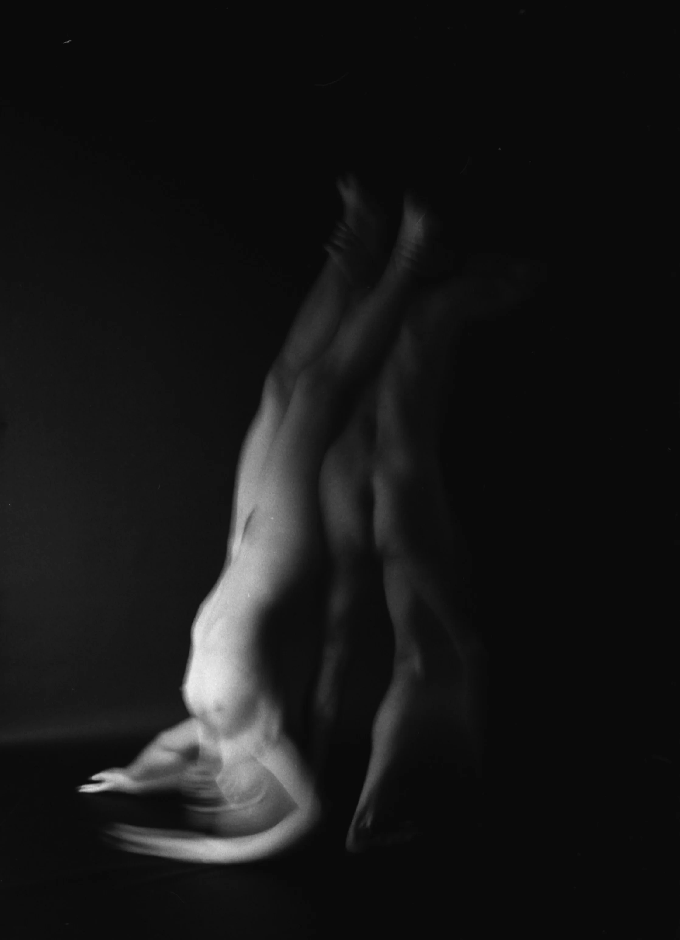 Black and white photo of a person with long hair, arms raised, with an abstract, blurry face and body, against a dark background.