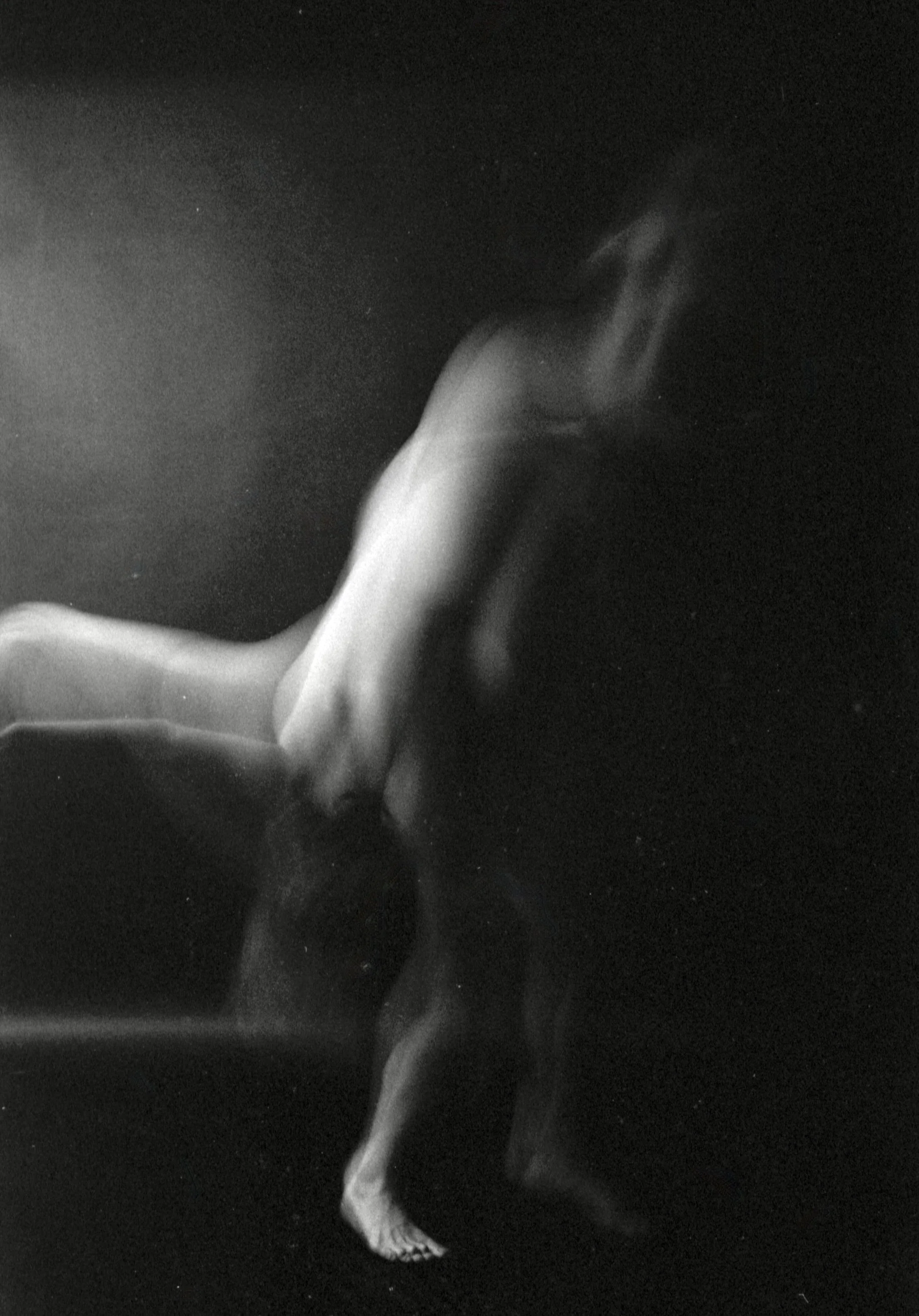 Black and white photo showing a person with one leg lifted and bent at the knee, their foot pointed downward, against a dark background.