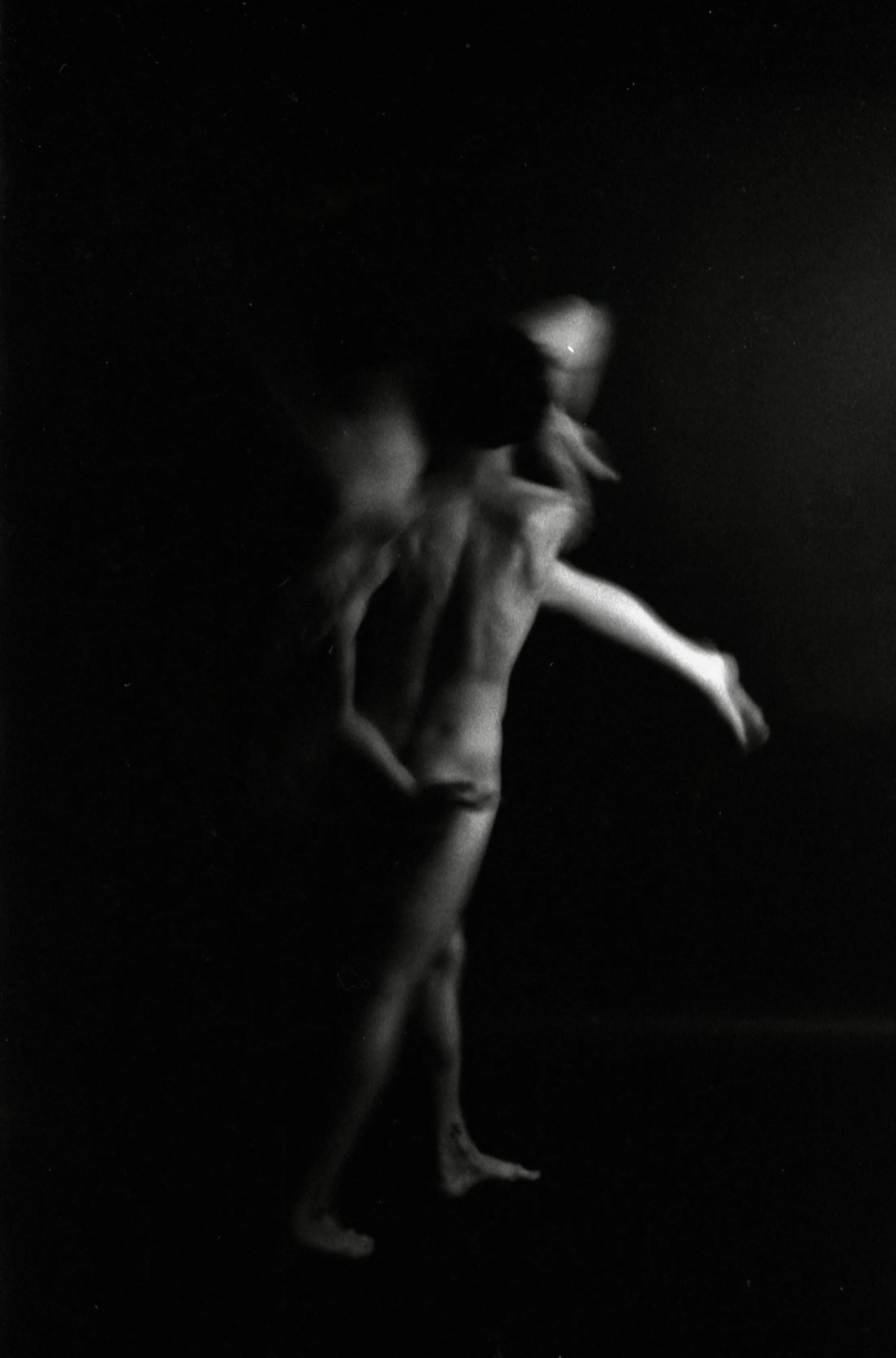 A black and white photo of a person with one leg extended and arms bent, against a dark background. The person’s face is partially lit, and their hair appears to be short.