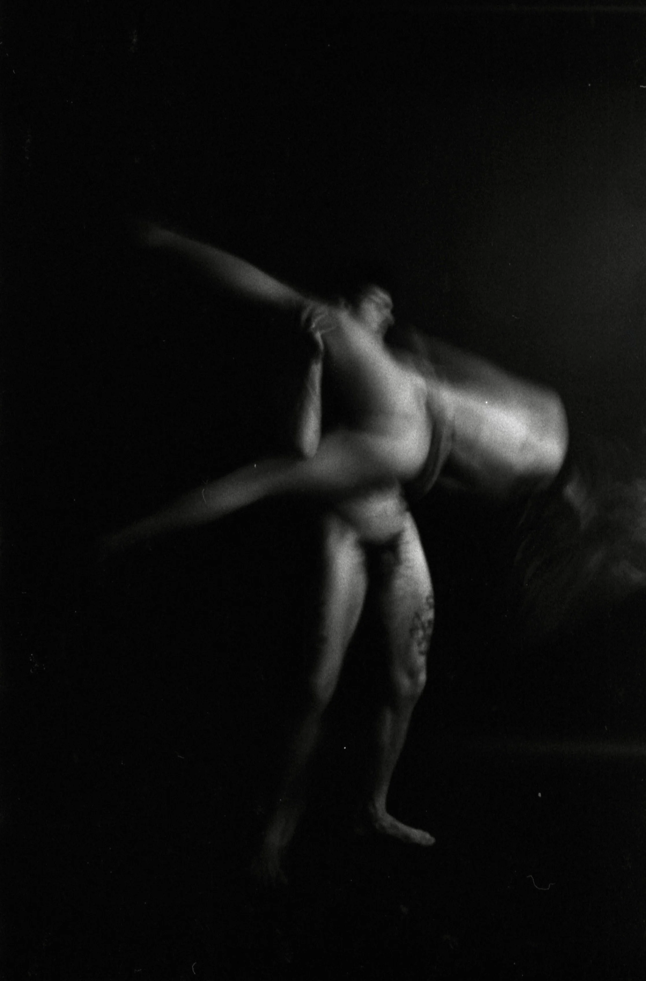 A black-and-white photo of a person with long hair, sitting in a fetal position with one arm covering their face, against a dark background.