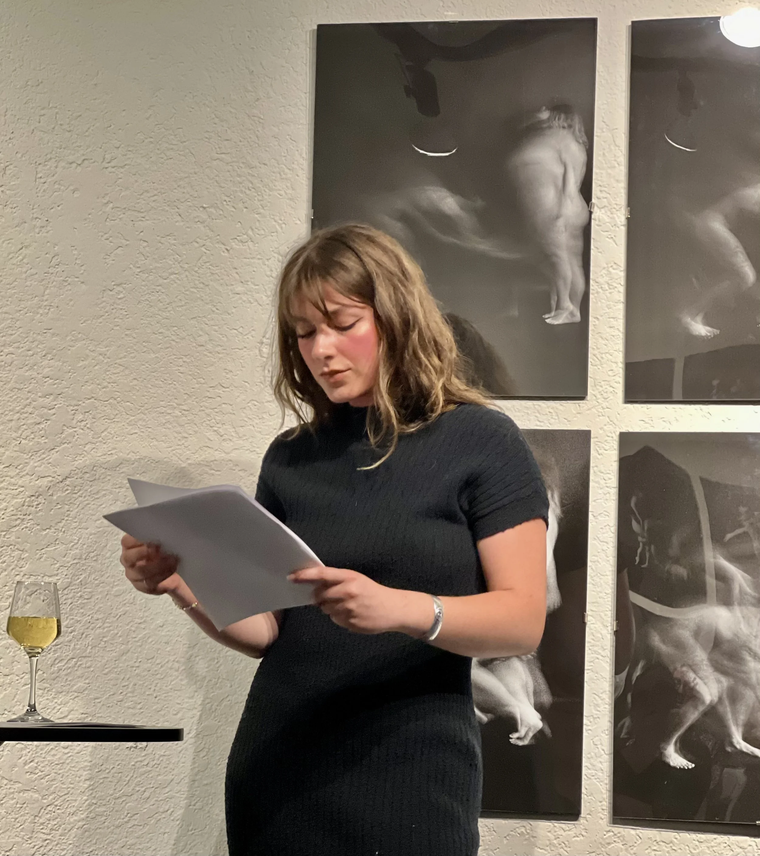 A woman with wavy brown hair wearing a black dress reading a paper in a room with beige textured walls and black and white art hanging on the wall. There is a glass of champagne on a small table to her left.