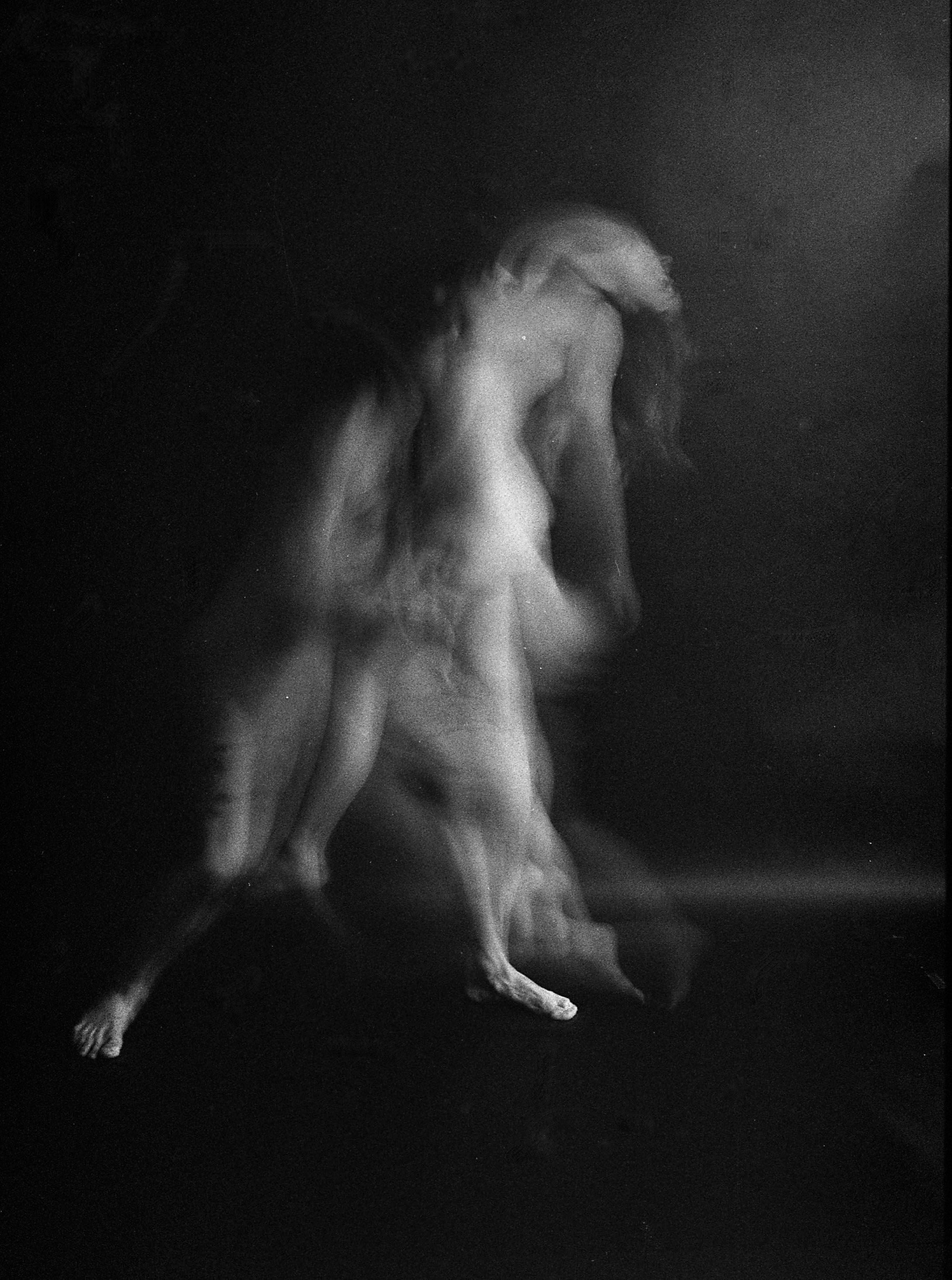 A black-and-white photo of a nude woman with long hair, facing downward, with one arm bent and hand on her knee, standing on a dark surface.