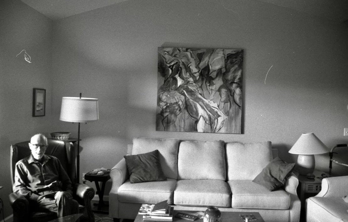 Living room with a man sitting in an armchair, a sofa with cushions, a painting on the wall, side tables with lamps, and a coffee table with books.