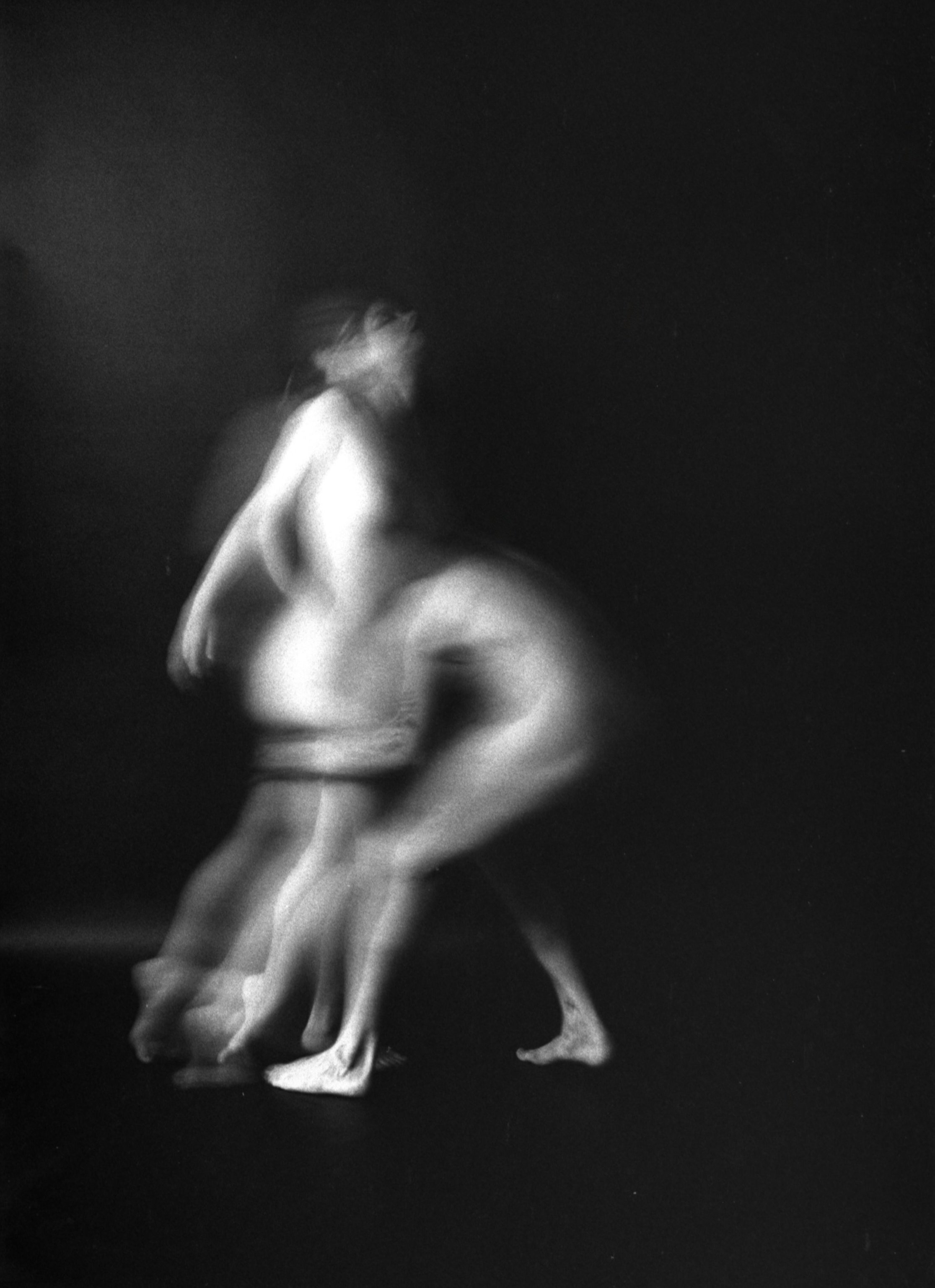 A blurred black and white photo of a person, possibly a woman, sitting or crouching on the ground, with a dark background.