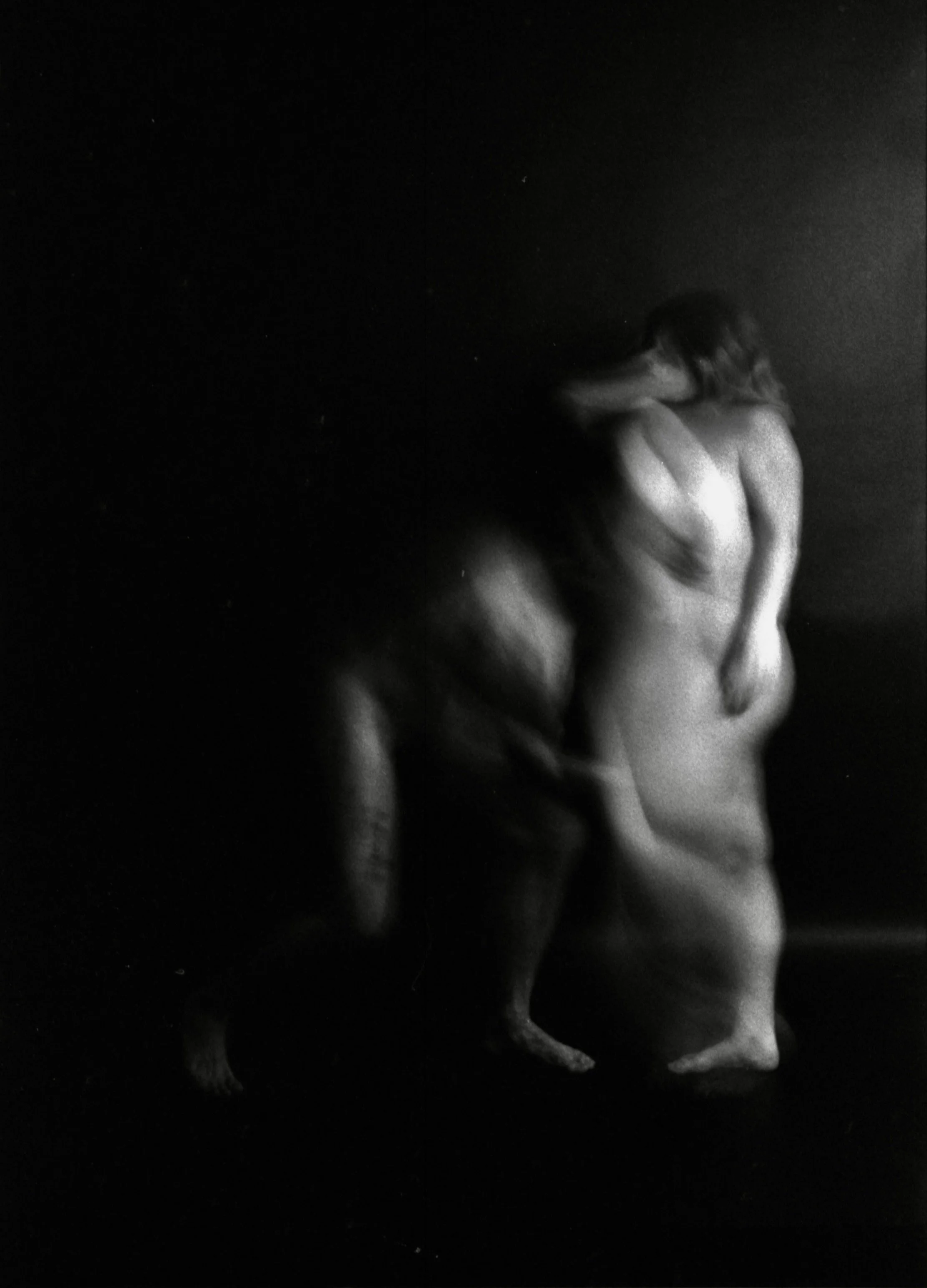A black and white photo of a nude woman sitting with her back turned, leaning forward with her face obscured by her hair.