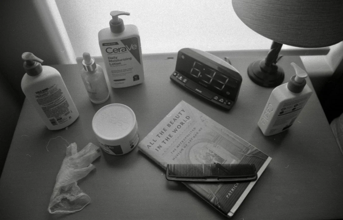A bedside table with personal care items, a clock showing 6:33, a magazine titled 'All the Beauty in the World,' and a comb with scissors.