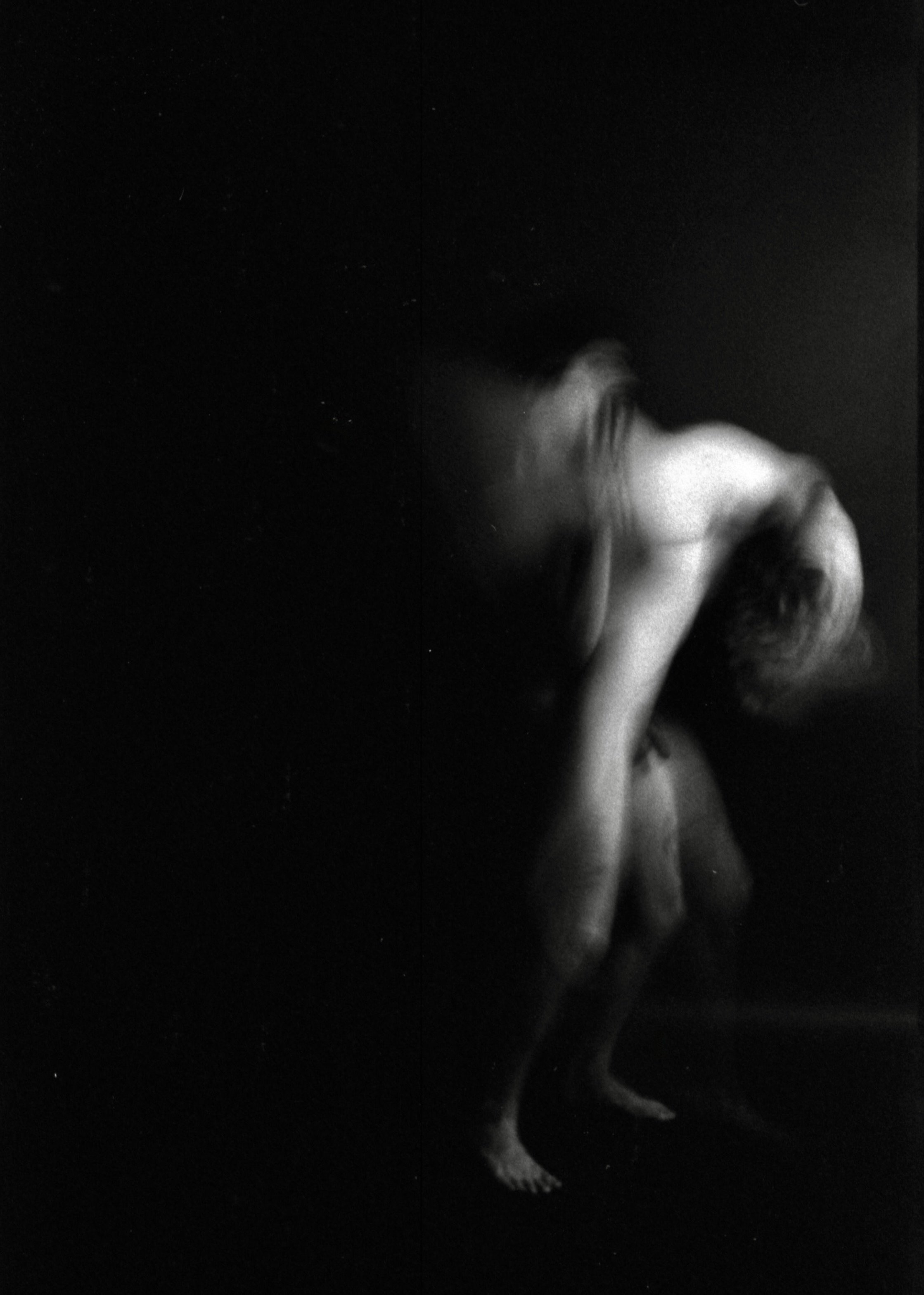 A black-and-white image of a person with short hair, sitting and holding their knees to their chest in a dark space.