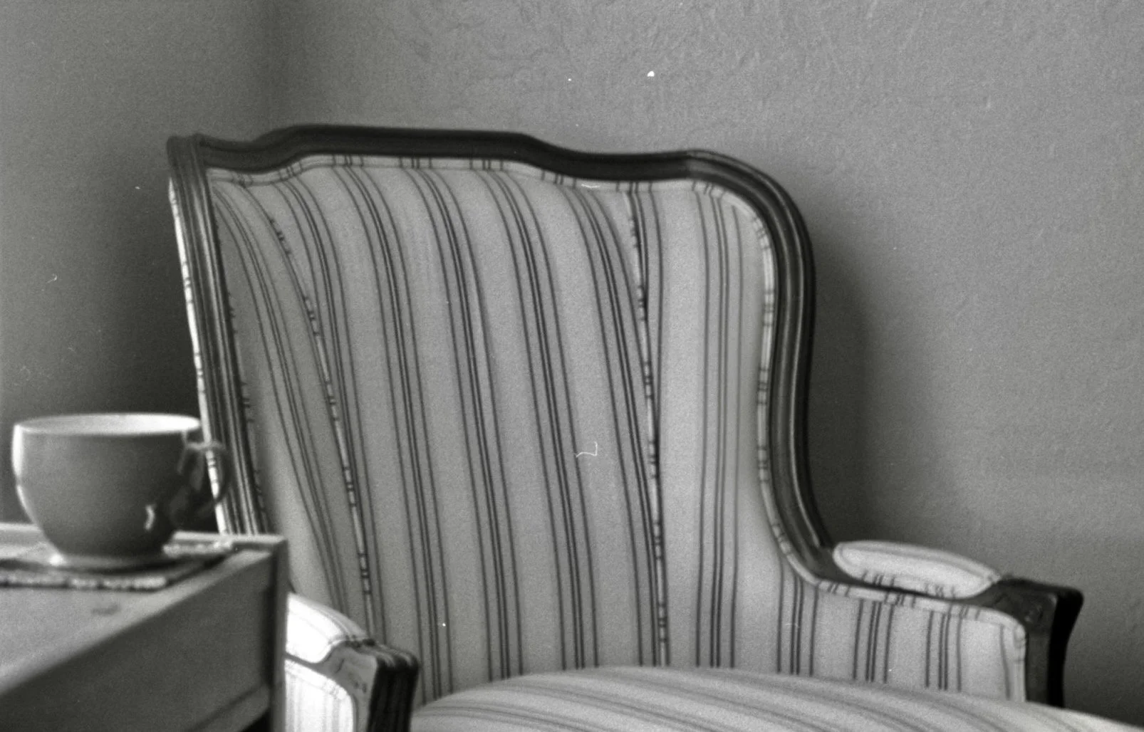 An empty vintage armchair with striped upholstery, placed against a plain wall, with part of a table and a cup visible in the foreground.