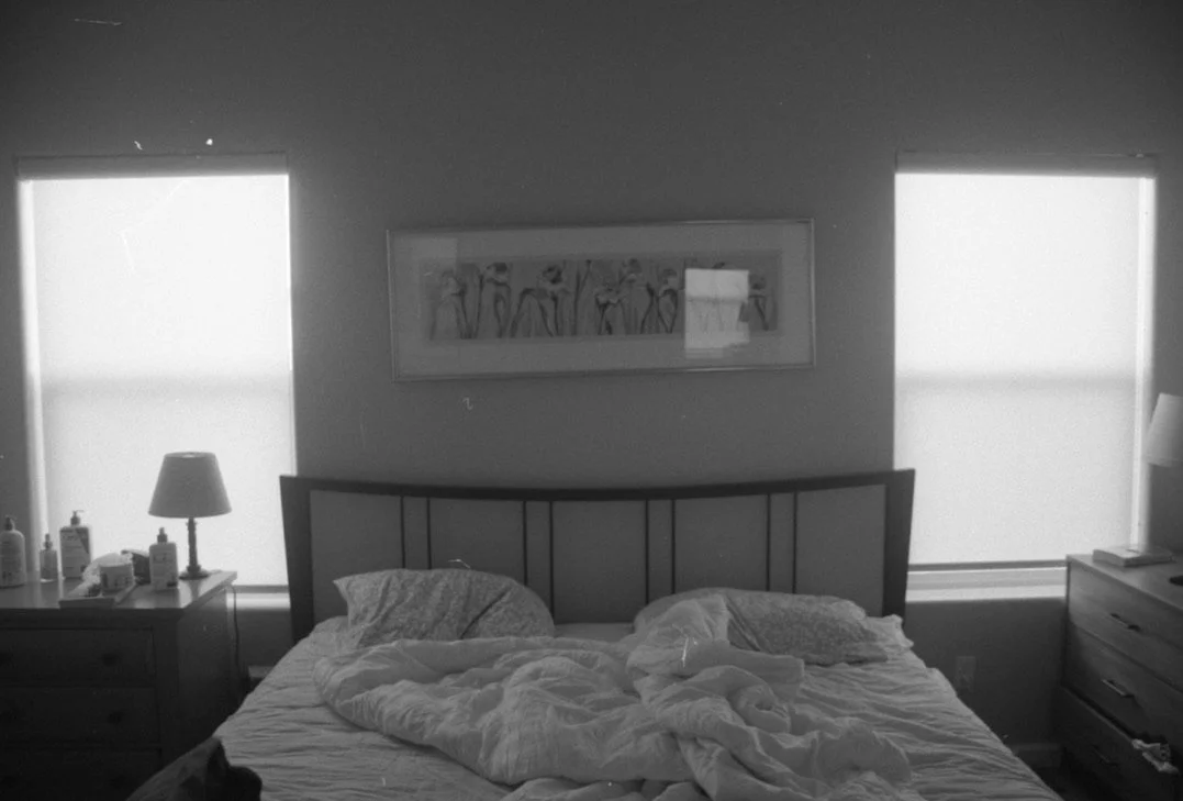 A bedroom with a messy bed, bedside tables with lamps, bottles, and other items, and two windows with blinds, in black and white.