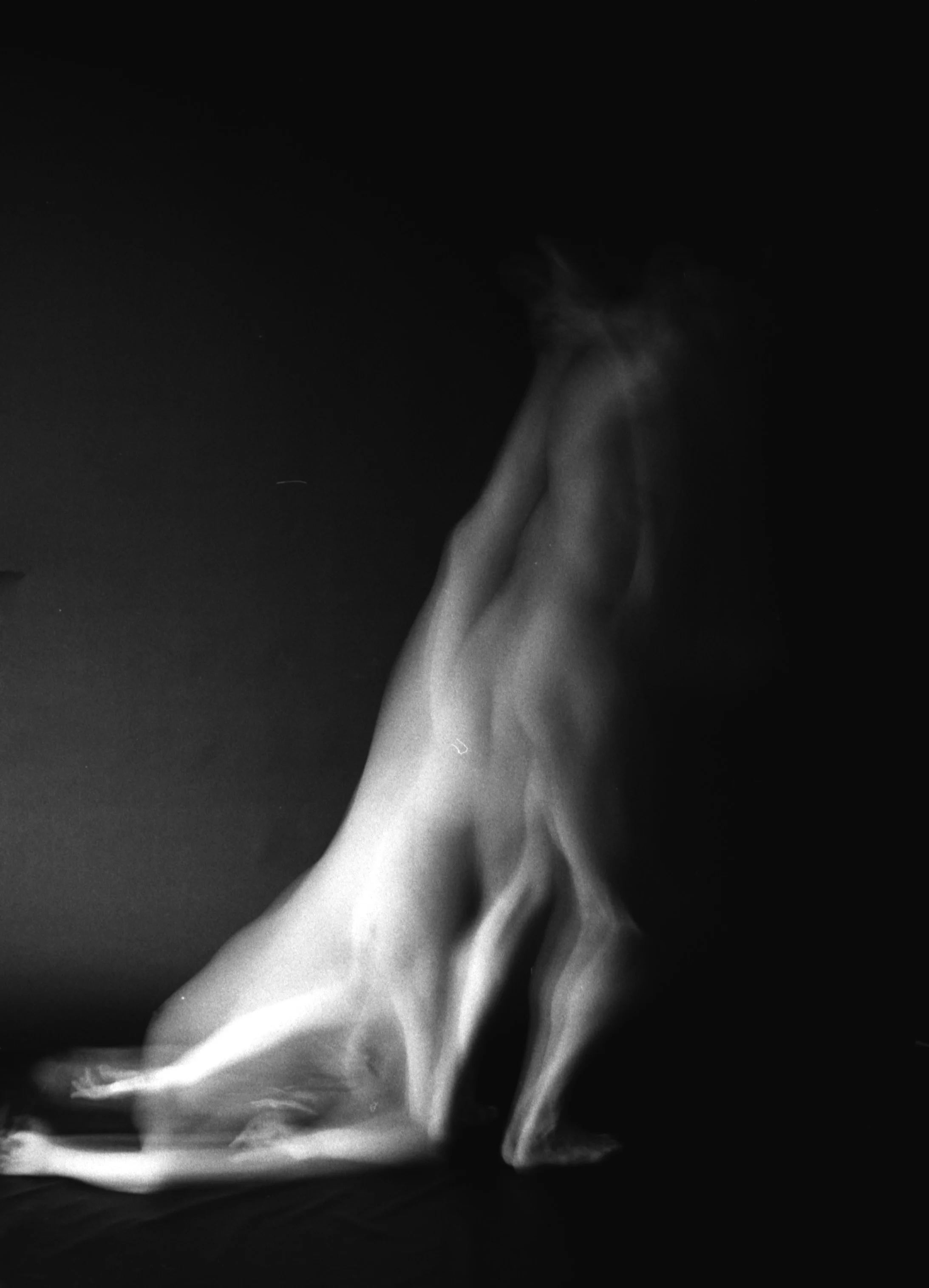 Black and white photograph of a person's lower legs and feet, with a flowing, draped fabric covering part of the legs. The background is dark and the lighting highlights the contours of the legs and fabric.