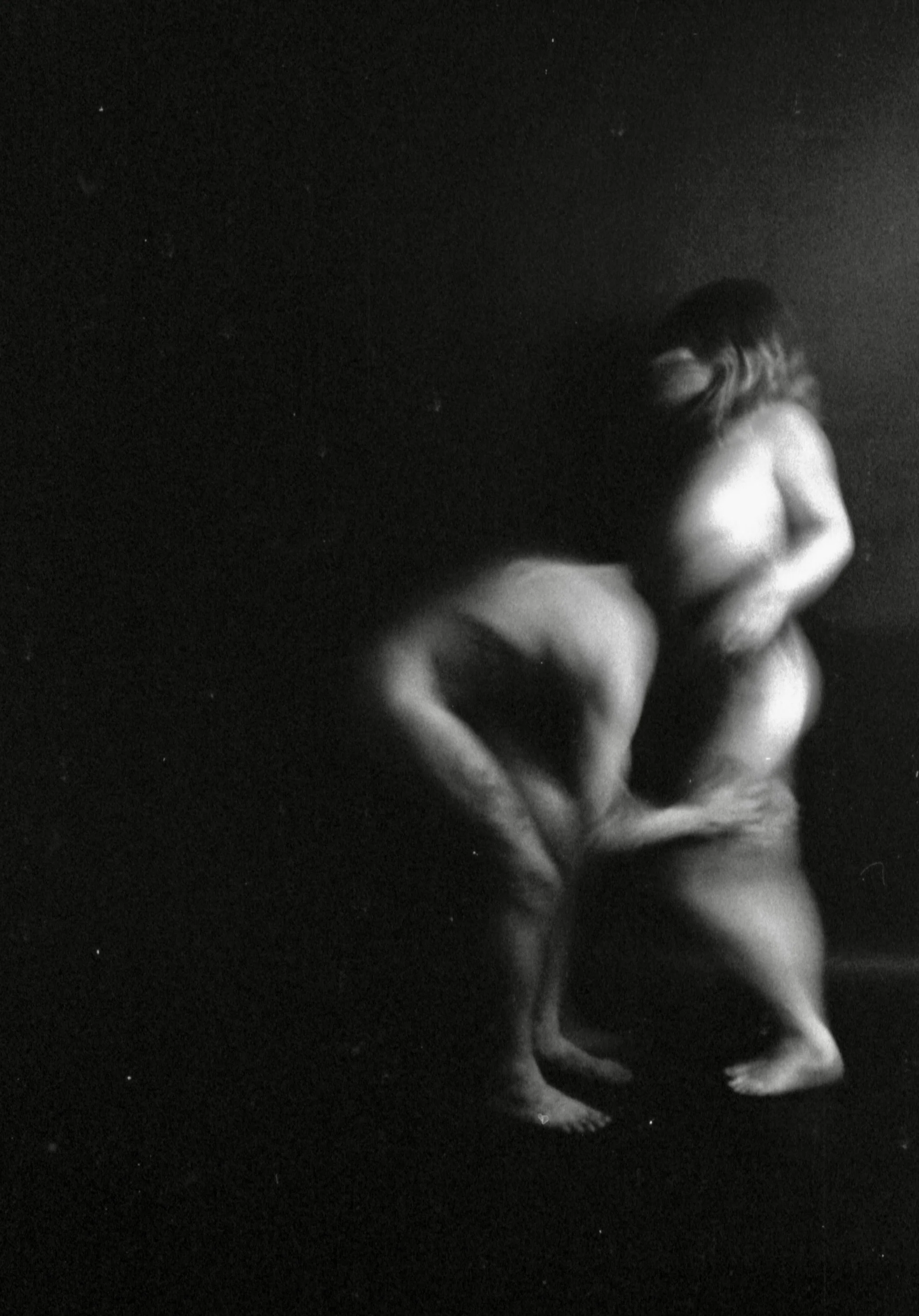 A black and white photograph of a nude woman crouching or sitting on the ground, with her head bowed and arms resting on her knees, set against a dark background.