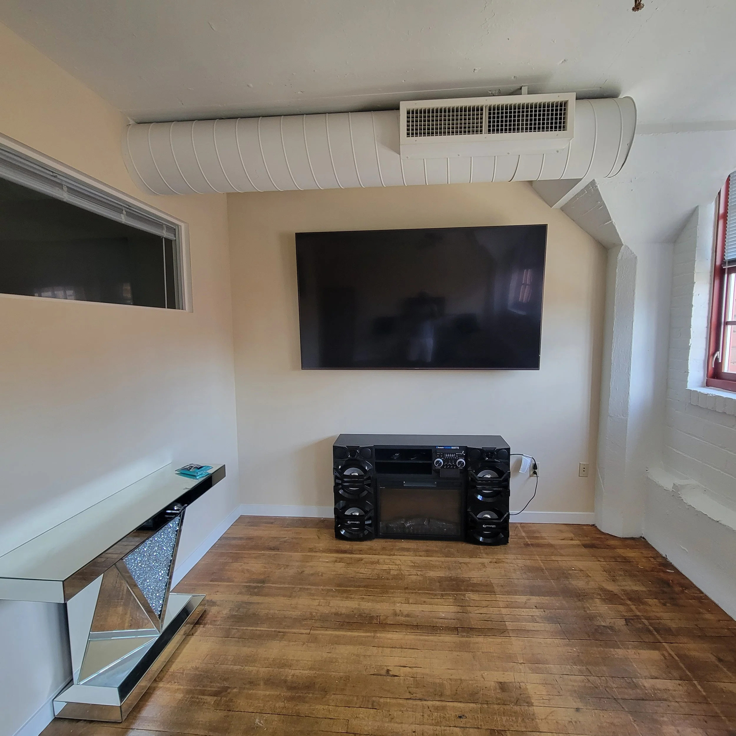 A living room corner with a wall-mounted flat-screen TV, a black audio system below it, a mirrored side table on the left, a window on the right, and a white air vent pipe running along the ceiling.