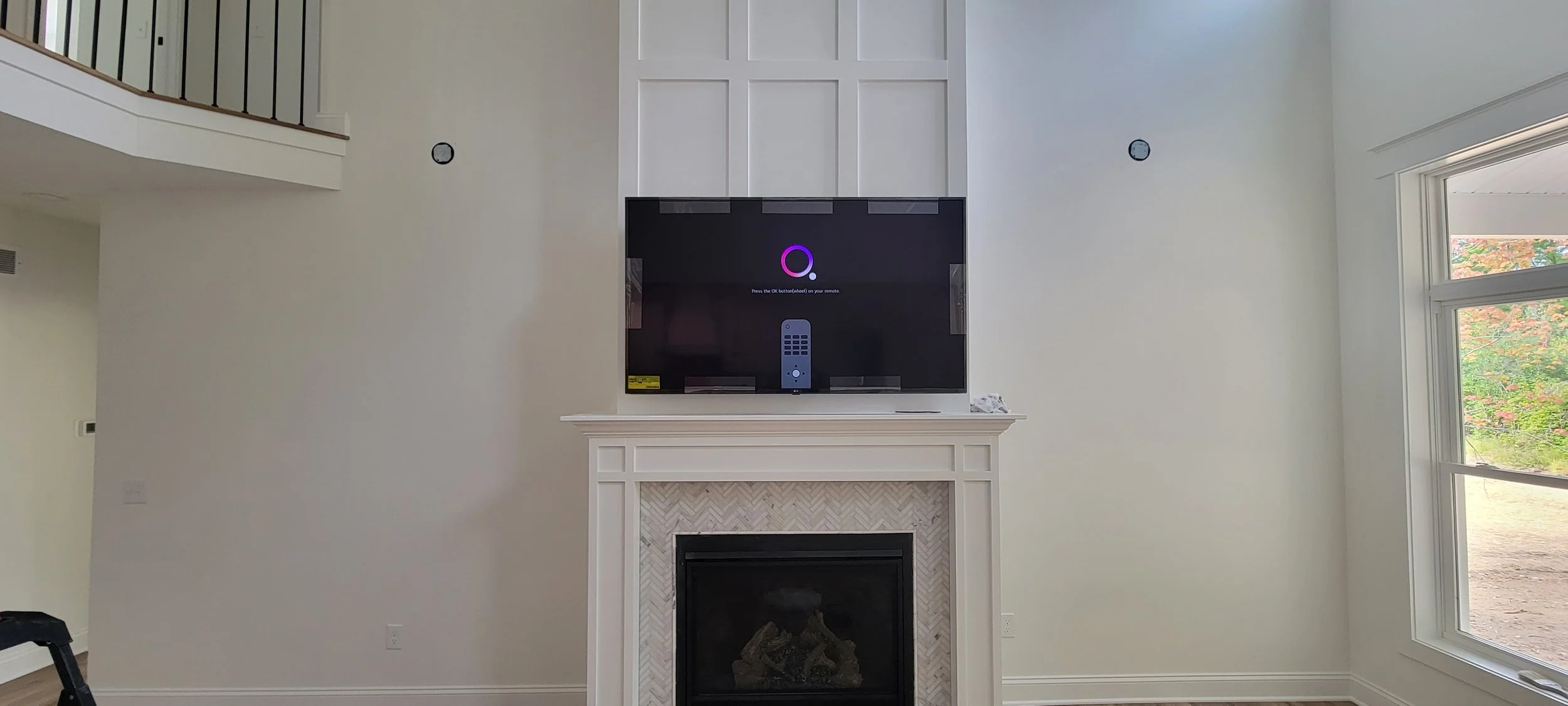 Living room with a fireplace, a television mounted above it, and a remote control on the mantle. The TV screen shows a setup process screen with a purple circle and remote control image, and there are large windows on the right side showing an outdoo
