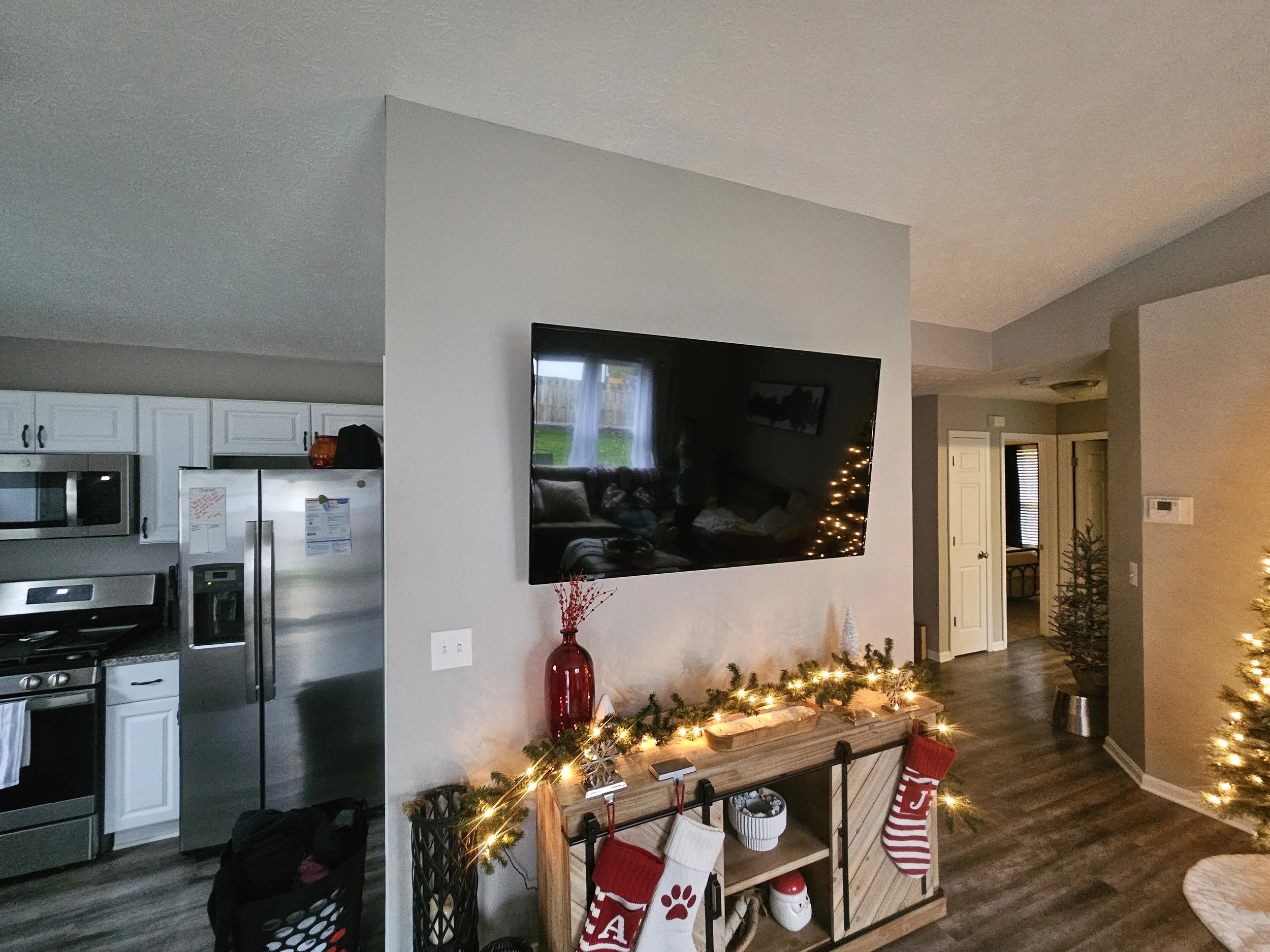 Living room with Christmas decorations, a large TV mounted on the wall, a console table with stockings, garland, and lights, and a Christmas tree to the right.