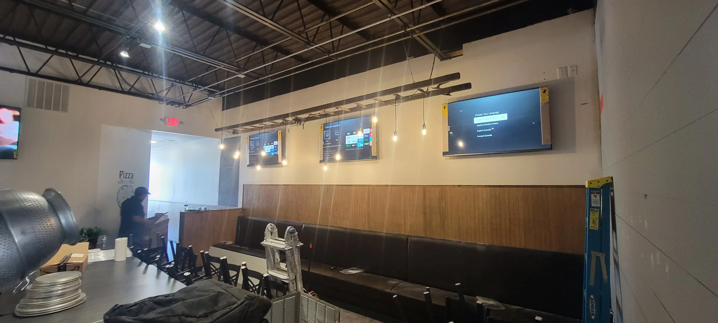 Interior of a restaurant or cafe with multiple television screens on the wall displaying a menu or interface, a long black cushioned bench along the wall, and a person standing near the counter. There are hanging light bulbs and some equipment or too