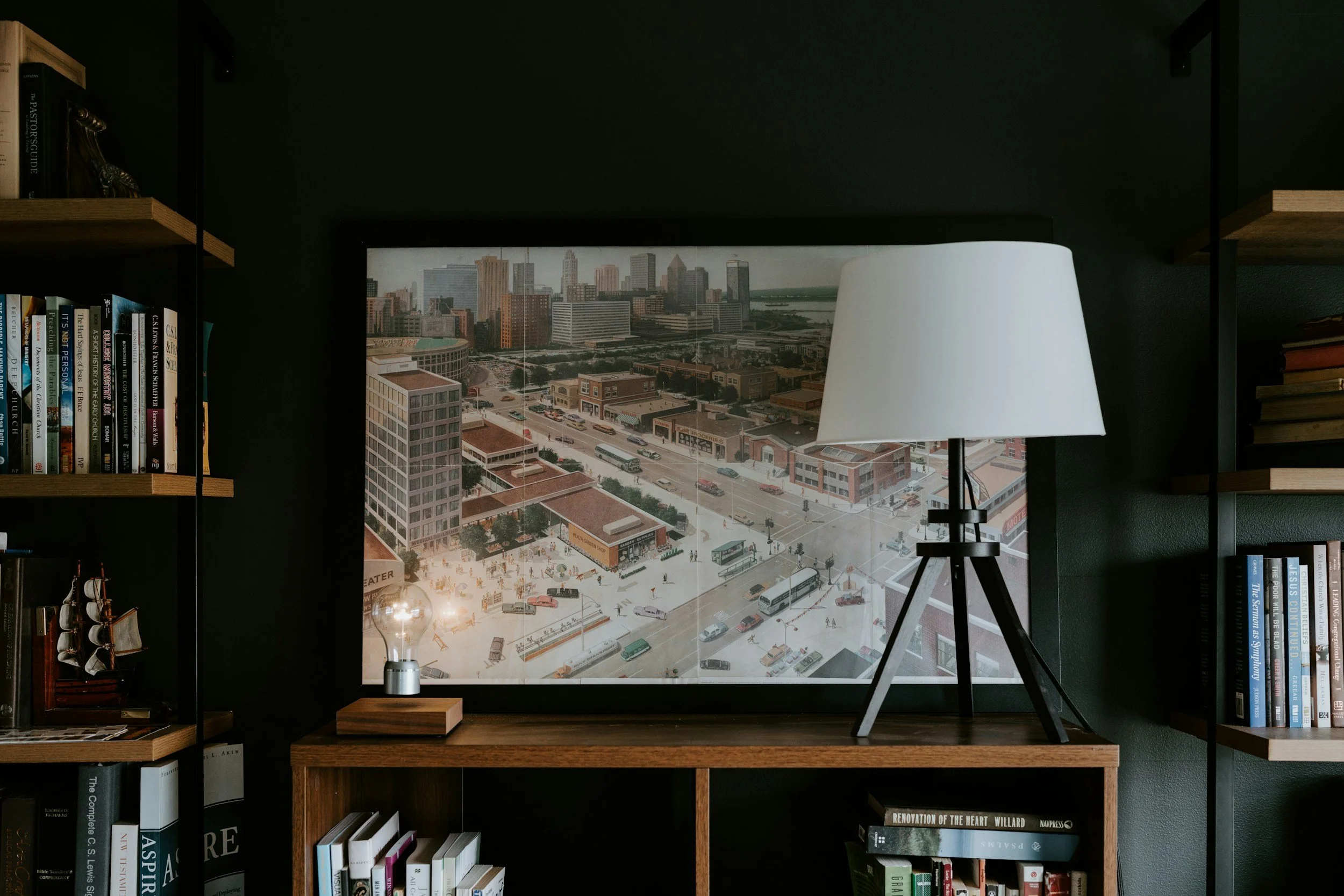 Interior of a room with a wooden bookshelf on both sides, a large framed cityscape photograph on a dark wall, a white tripod table lamp, and a small decorative ship on the left shelf.