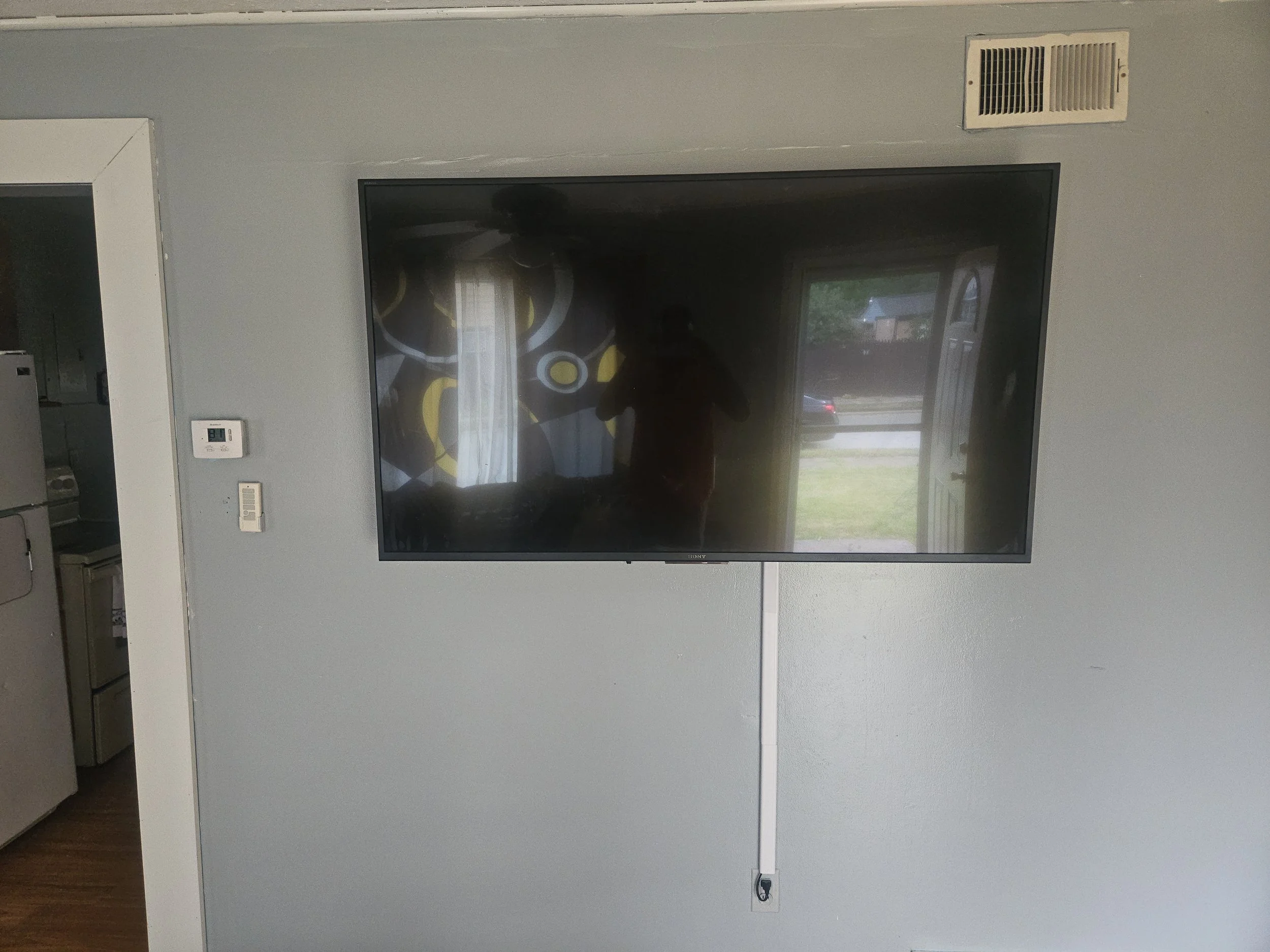 A flat-screen TV mounted on a gray wall in a living room, with a white cable hanging down from the bottom of the TV. The reflection on the screen shows a person taking a photo, a window with curtains, and a door opening to an outside view.