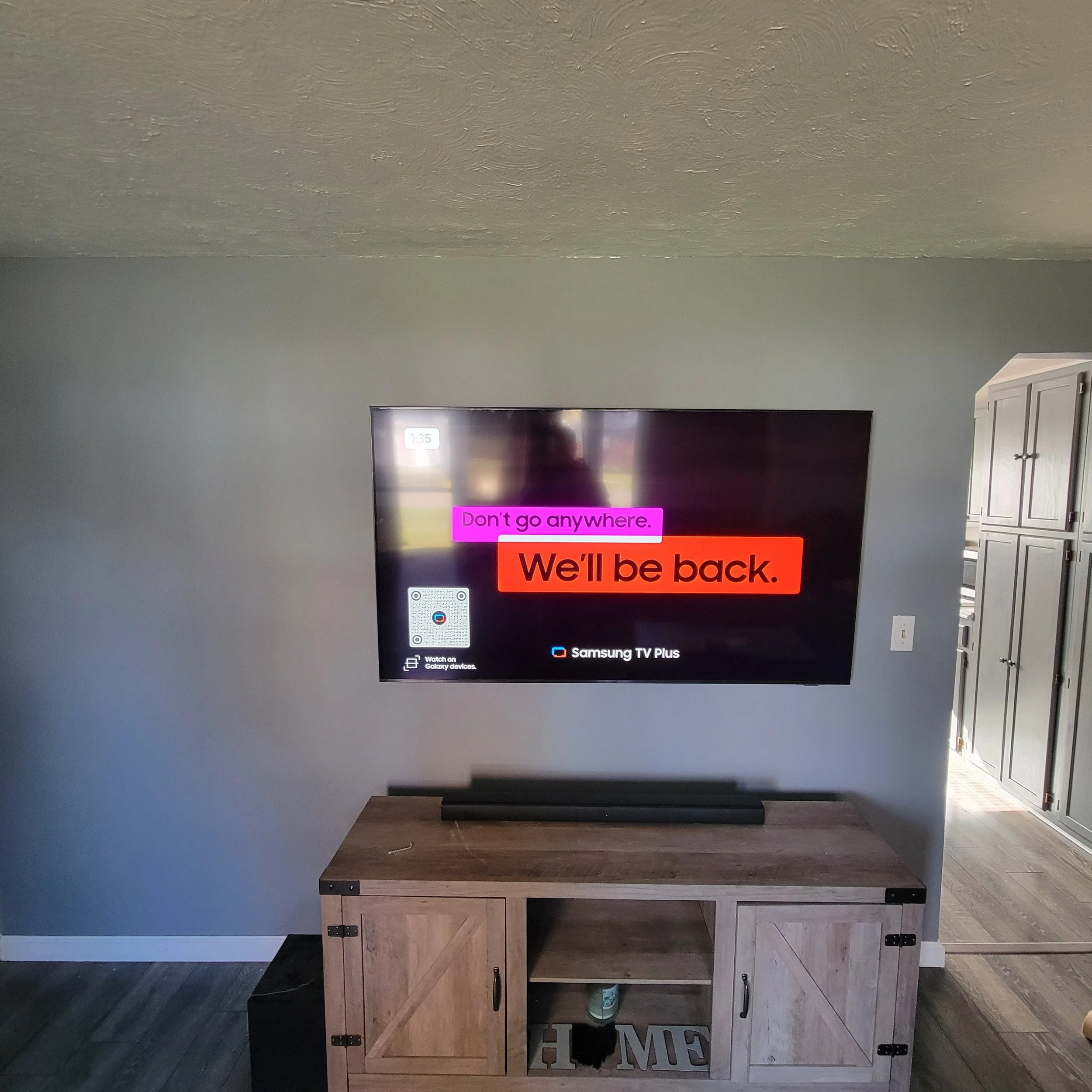 TV screen displaying message 'Don't go anywhere. We'll be back.' with a Samsung TV Plus logo, mounted on a gray wall above a wooden credenza, in a living room.
