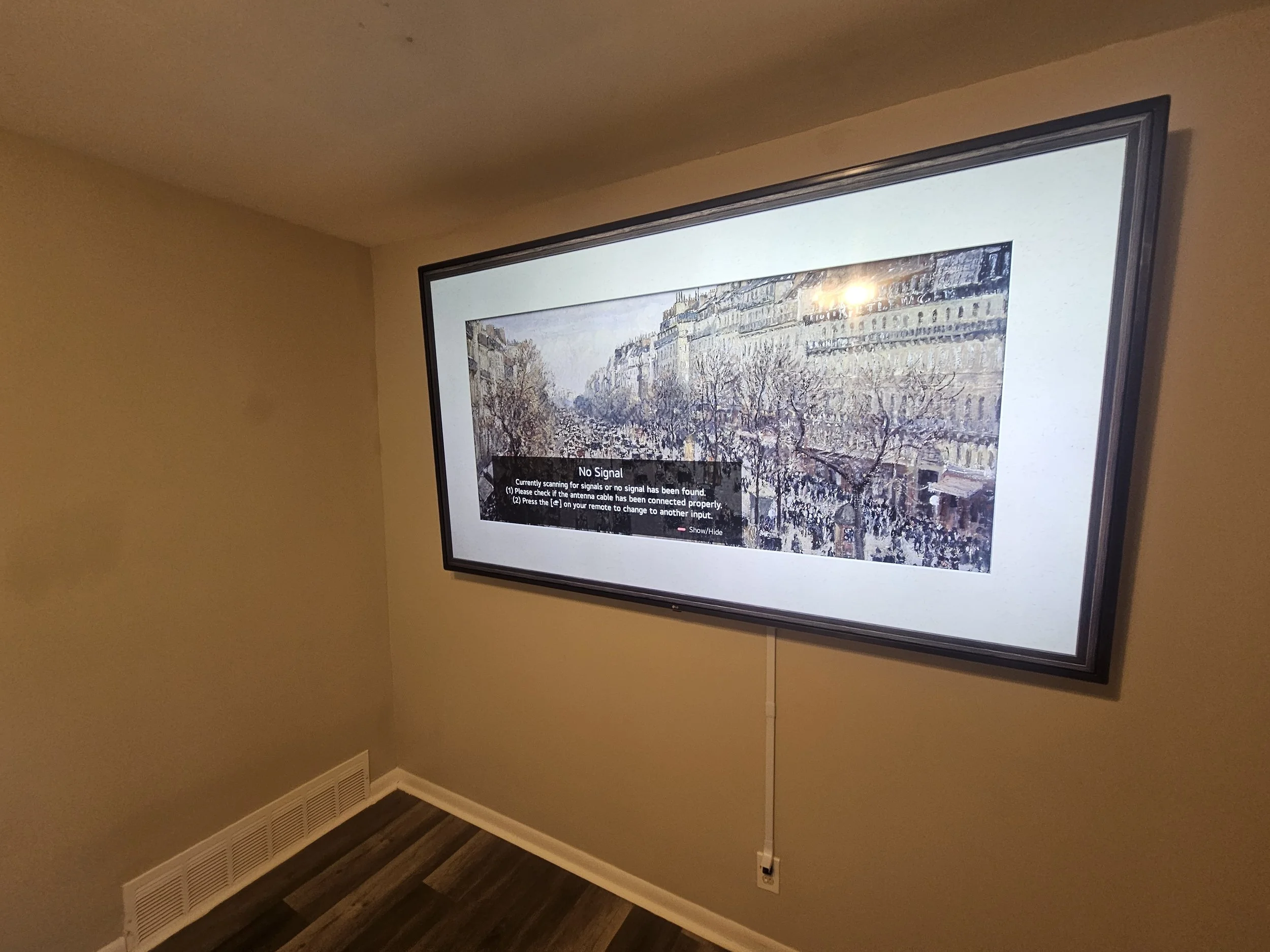 A large flat-screen TV mounted on a beige wall displays a painting of a street scene with trees and buildings, with a black notification box in the center reading 'No Signal' and instructions for checking the antenna cable.