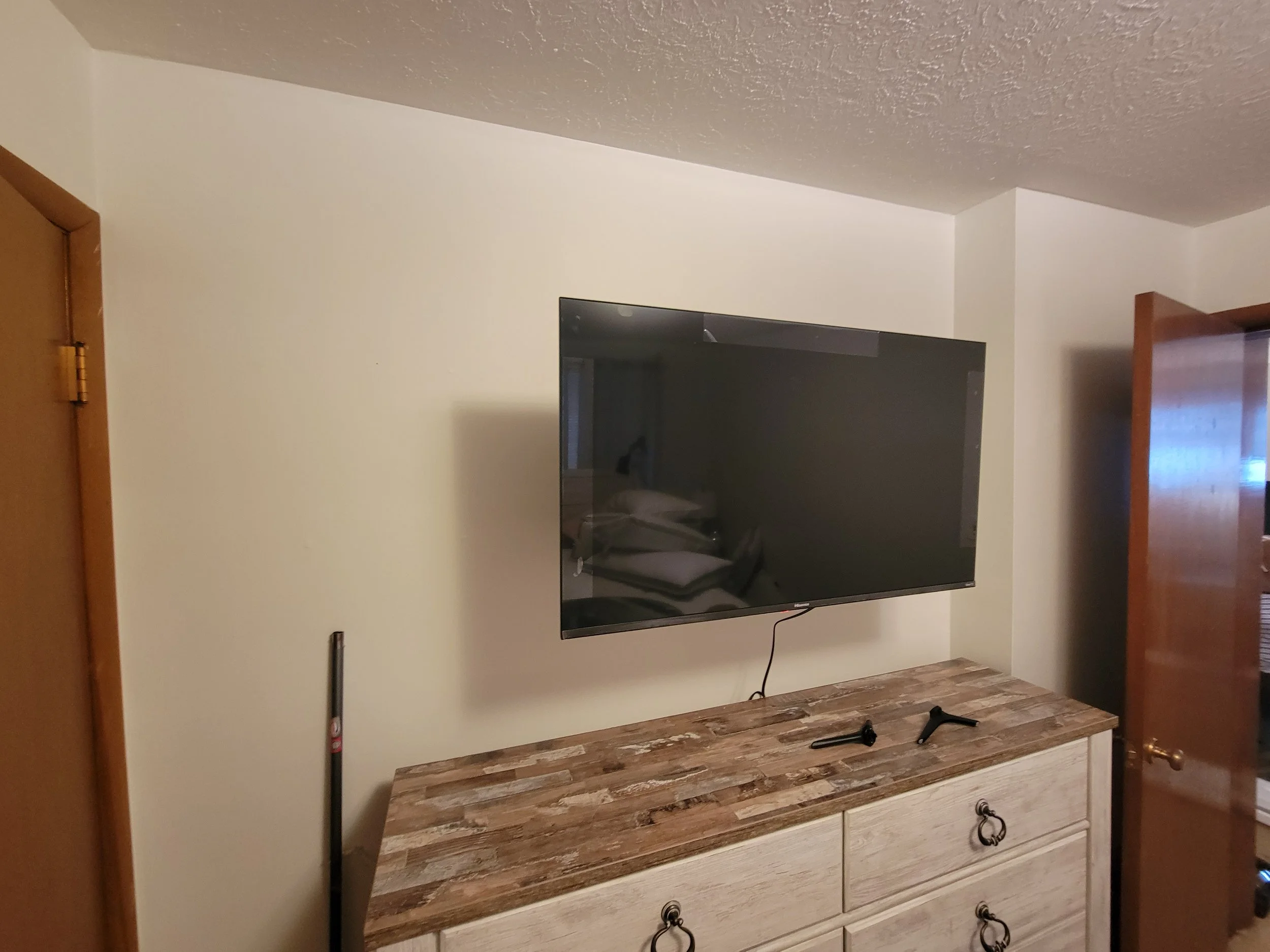 TV mounted on a wall above a wooden dresser with two black tools on top, next to a closed door.