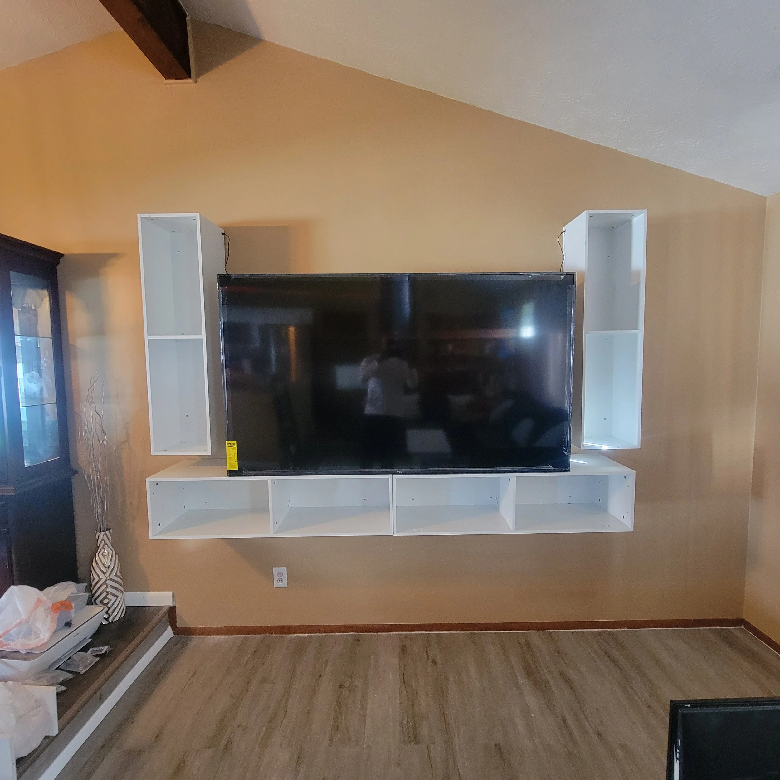 Empty white wooden shelves surround a large flat-screen TV mounted on a yellow wall inside a living room.