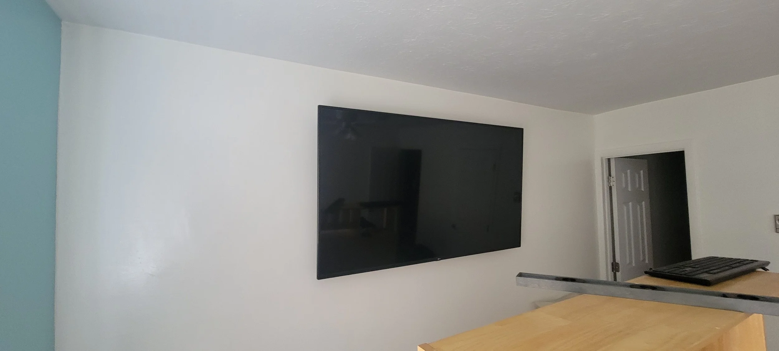 A plain room with white walls and a wall-mounted flat-screen television. There is a doorway with a white door, and a countertop with a keyboard and some objects on it.